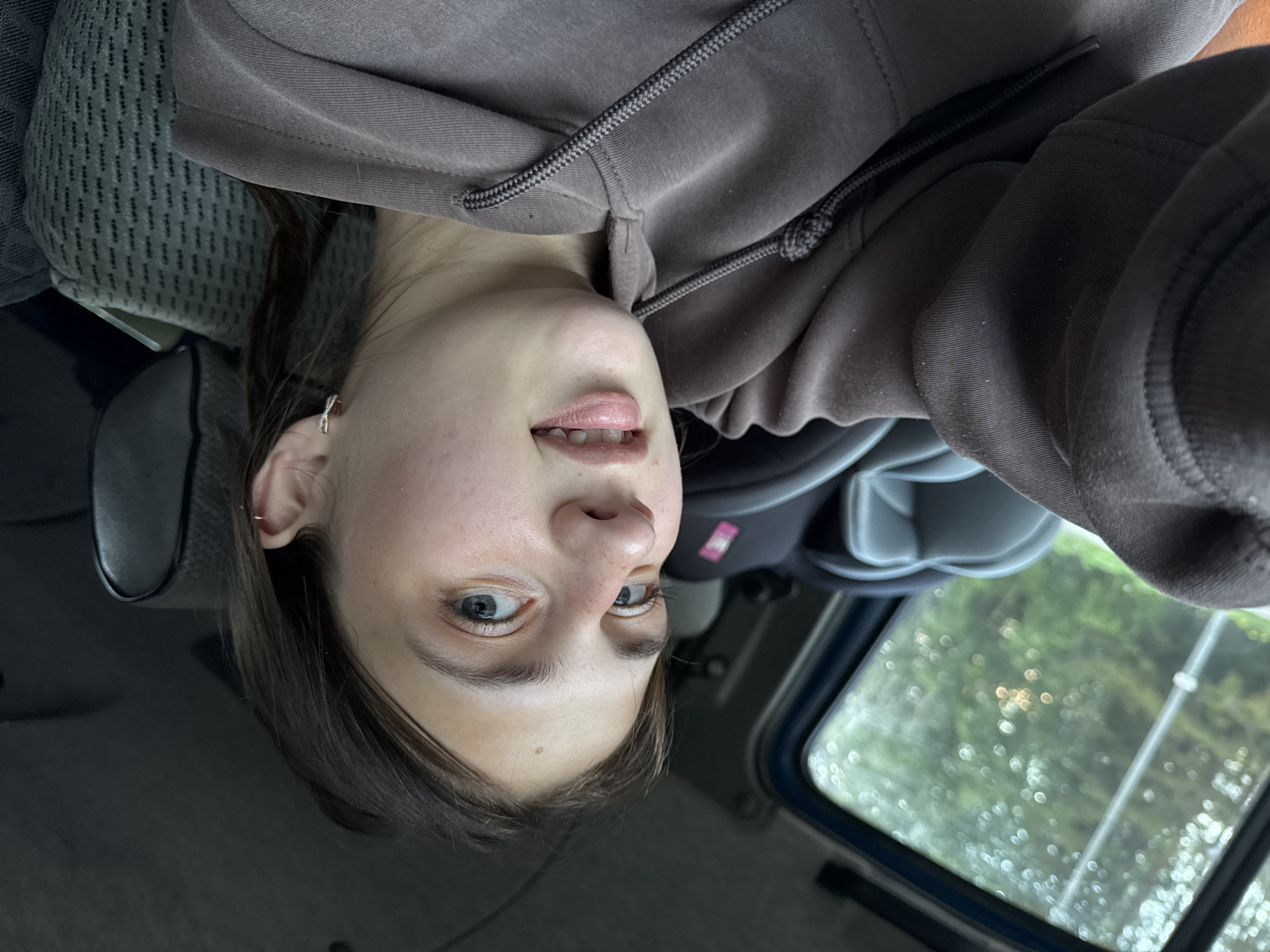 A young woman with light brown hair and blue eyes is seated in the passenger seat of a vehicle. She is wearing a brown hooded sweatshirt. Her left arm is extended, and she is holding the camera to take a selfie. The background shows the interior of the car, including a car seat and the car window, which looks out onto a blurry, green, tree-filled landscape. The weather outside appears to be overcast, possibly raining, given the bokeh effect on the foliage. The image does not provide any specific details about the location in the city of Vary, Russia.