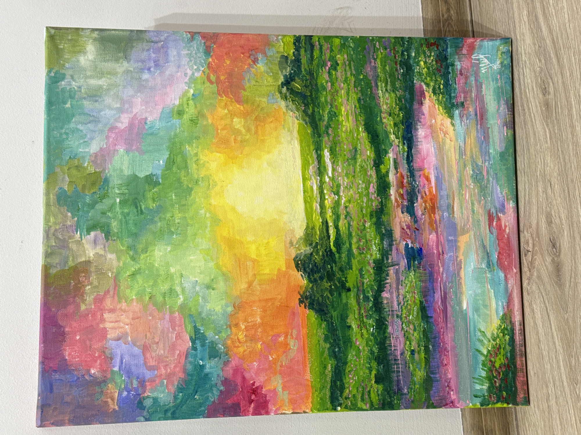 An abstract painting on canvas is displayed. The painting features a blend of colors including green, yellow, orange, pink, purple, and blue. Vertical brushstrokes create a sense of depth and texture. The painting is positioned against a white wall on the left side and a wooden surface on the right side. No people or discernible objects are visible in the background. The painting's style suggests a natural scene, possibly a forest or garden, but it is not clearly defined. There is no indication of it being located in Yerevan, Armenia.