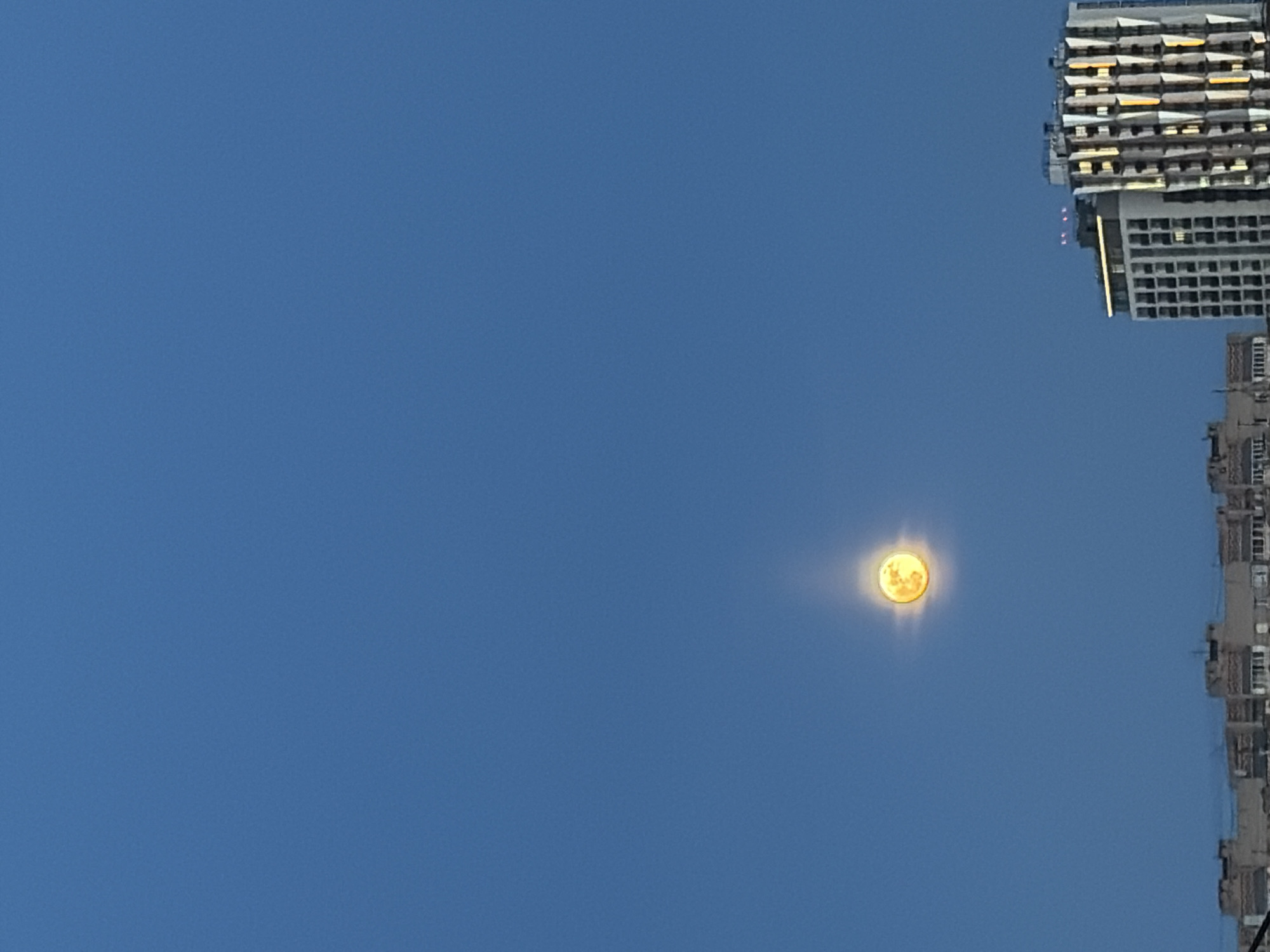A low-angle, outdoor shot shows a clear blue sky with a bright yellow object that may be the sun or moon. In the upper right corner, the top of a tall building is visible, appearing sideways. To the right of the bright object and closer to the bottom of the frame, part of another building is visible, also sideways. There are no people visible. There is not enough information in this image to determine its location in Izhevsk, Russia.