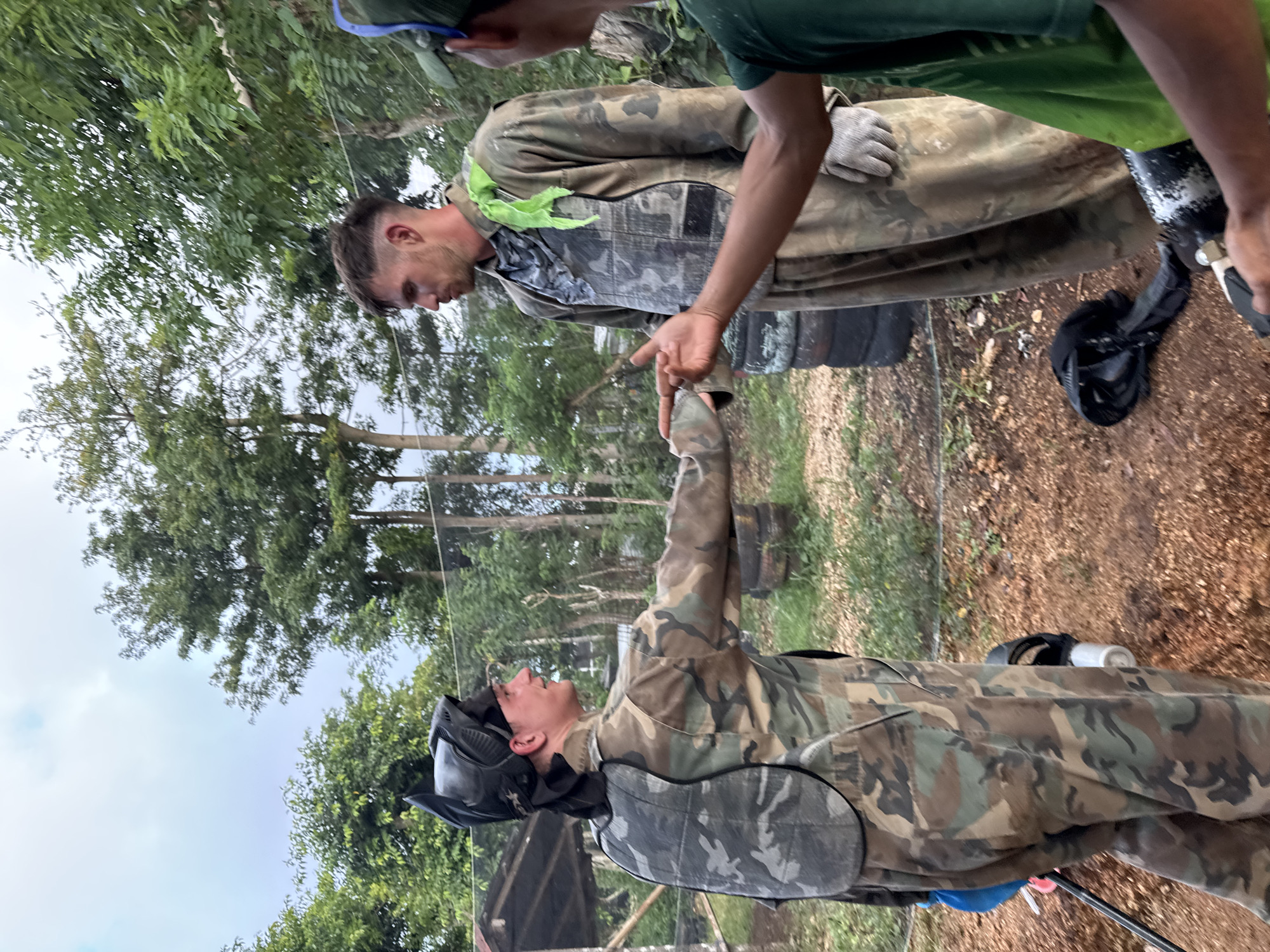 A group of men, seemingly in a paintball arena, are interacting in an outdoor setting. Two men are dressed in camouflage gear, with one wearing a paintball mask and extending his hand for a handshake. Another man, also in camouflage, extends his hand to meet the first's. A third man, wearing a green shirt, is positioned above them. The ground is covered in gravel. The location appears to be a paintball facility with tires visible in the background, and trees visible in the distance. The city of Kuta Selatan is not discernible from the image.
