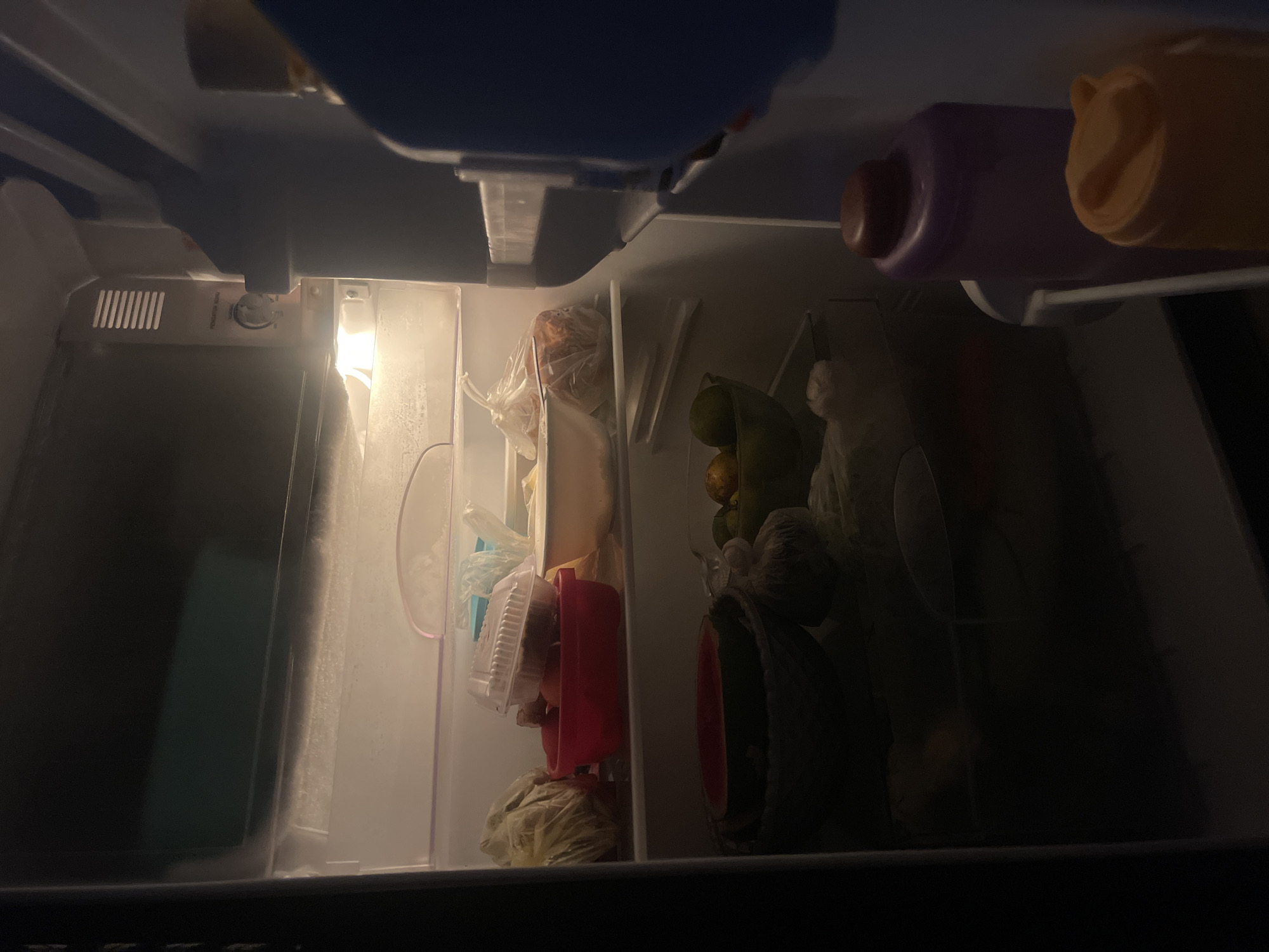 A dimly lit refrigerator is filled with various food items and containers. On the top shelf, a plastic container with food sits on top of a red bowl containing more food. To the left, a plastic bag holds produce. On the shelf below, a large bowl contains what appears to be a watermelon, and next to it, a green bowl holds citrus fruits. In the foreground, two large bottles, one purple and one orange, are visible on the door. The refrigerator is located in the city of Anggaswangi, Indonesia.