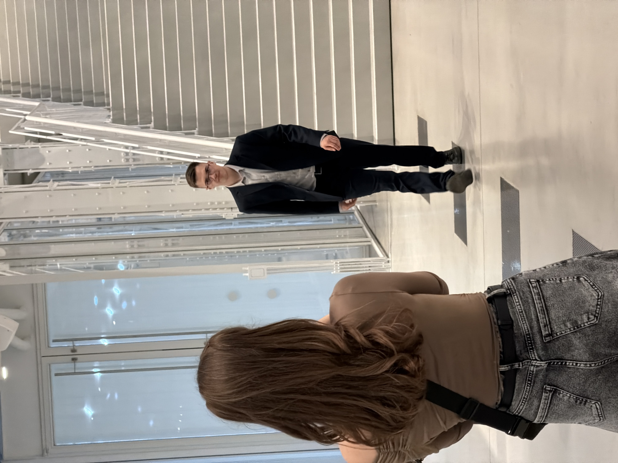 A man in a suit stands near a staircase while a woman with long brown hair faces away from the camera. The scene takes place indoors, likely in a modern building. Based on the architecture of the staircase and windows, the location could be in Moscow, Russia.