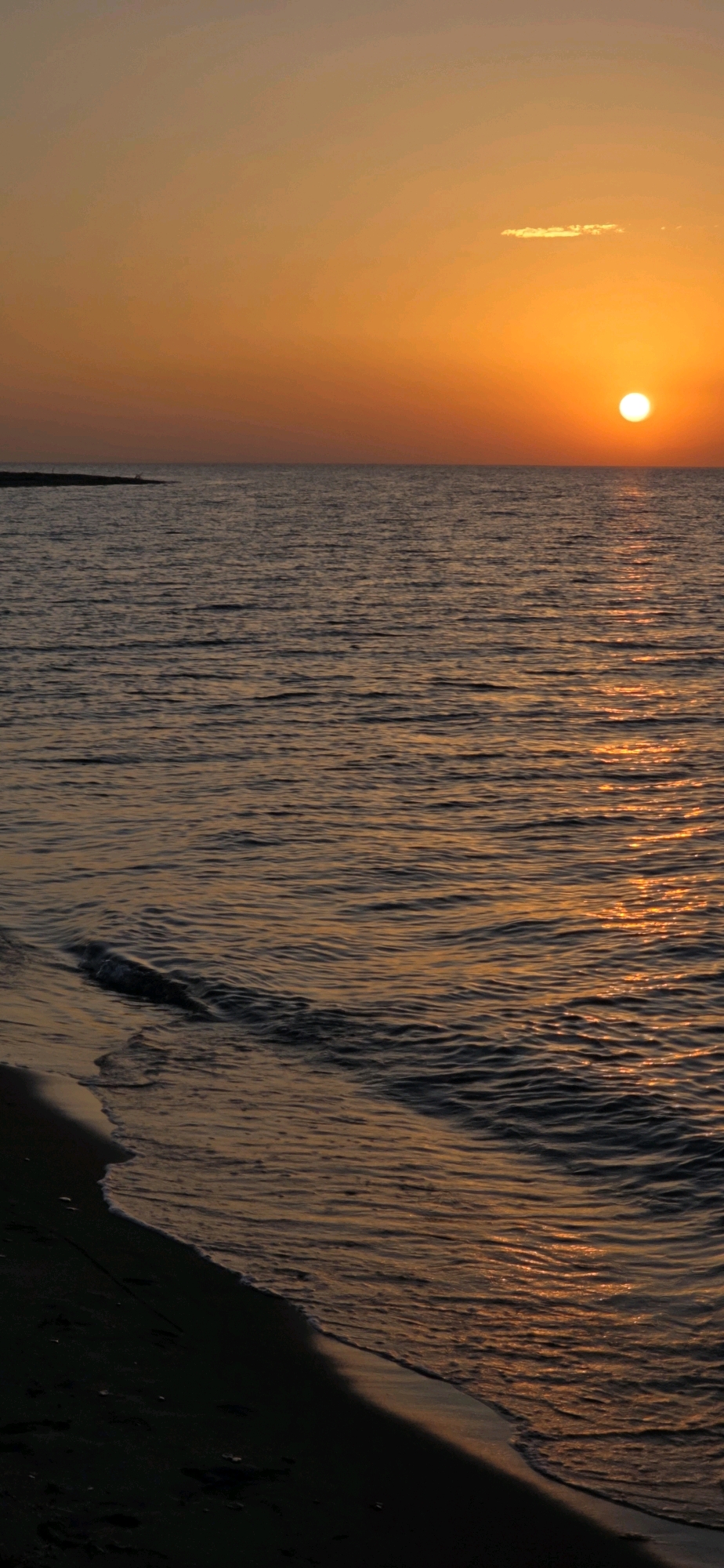 The image depicts a serene beach scene at sunset in Basbeshik, Kyrgyzstan. The sun, a vibrant orange orb, is low on the horizon, casting a warm glow across the sky and the rippling water. The sea is calm, with gentle waves lapping at the dark, sandy shore. The water reflects the colors of the sunset, creating a shimmering path of light from the sun to the foreground. A small, dark landmass is visible in the distance on the left side of the image. No people are visible in the scene, and no actions or interactions are taking place. The environment is natural and peaceful, characterized by the interplay of light, water, and land.