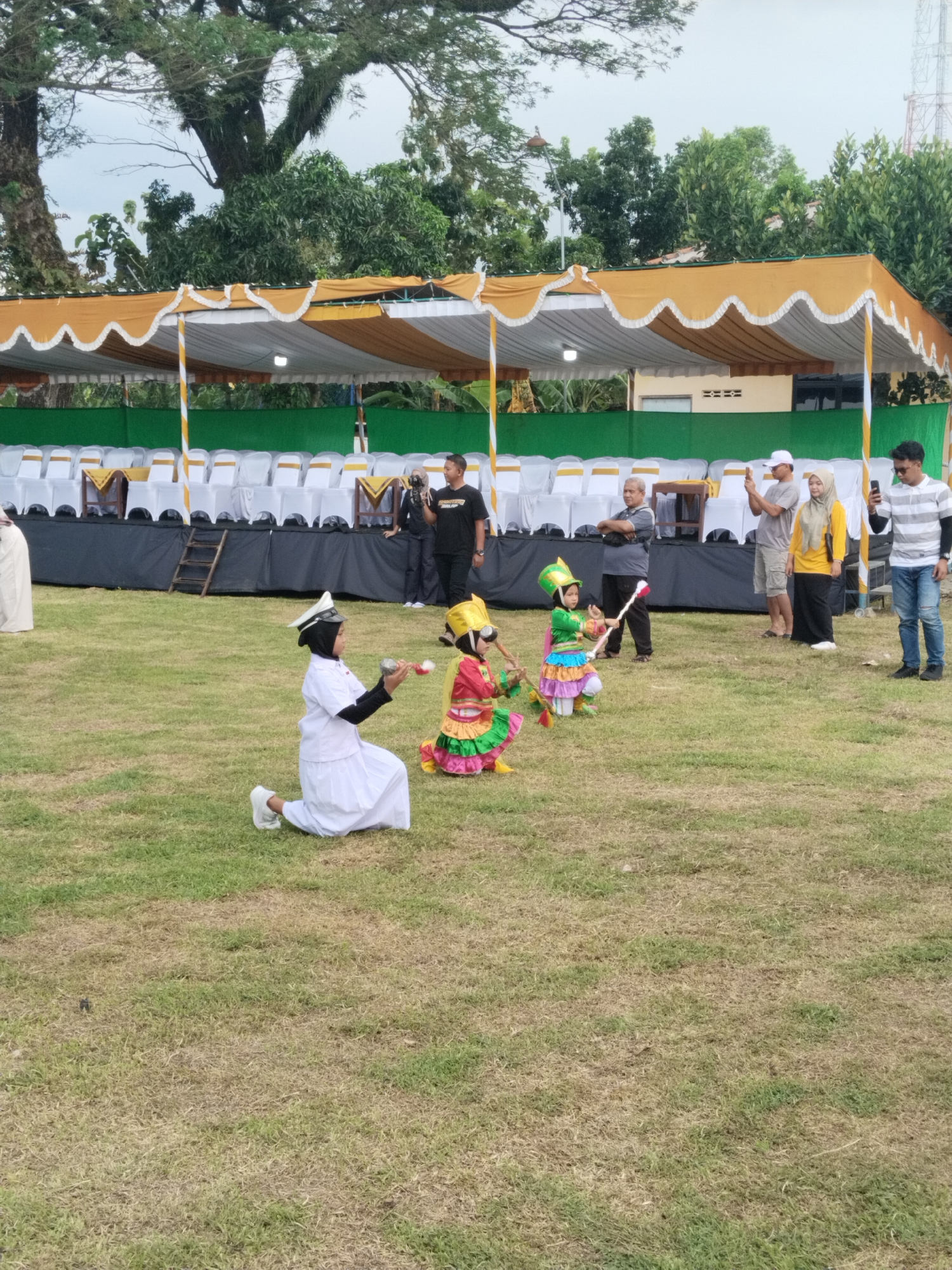 The image captures an outdoor scene in Sambirejo, Indonesia, featuring a grassy field where three children are engaged in a performance.
One child, kneeling in a white uniform and a black headscarf with a white cap, holds small batons. Two other children, dressed in bright traditional costumes with conical hats, are also kneeling and manipulating batons.
Behind the children, several adults are watching the performance, some capturing the moment with their mobile phones. A covered stage area is visible in the background, with rows of white chairs and a ladder. Trees and a telecommunications tower are also in the background.