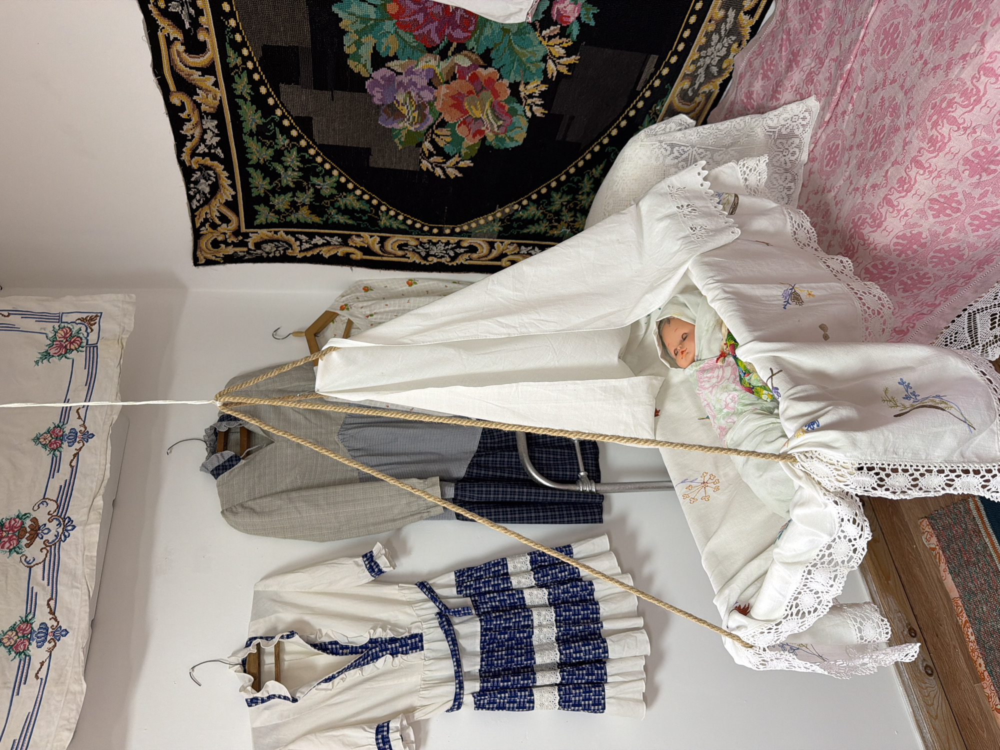 The image depicts a room, possibly in Ust'-Kabyrza, Russia, designed to resemble a historical nursery. In the center, a white, fabric-lined bassinet is suspended by ropes, containing a doll dressed in a light-colored outfit with floral patterns. The bassinet is adorned with white lace trim and embroidered details. Surrounding the bassinet, several pieces of clothing are displayed on hangers against a white wall. These include a white dress with blue trim and a plaid pattern, a grey and blue checkered dress, and another white dress with small floral embroidery. A large, dark tapestry with vibrant floral motifs hangs on the right wall. A pink patterned fabric covers a surface to the right of the bassinet, suggesting a bed or a similar piece of furniture. The overall impression is of a staged exhibit, showcasing traditional clothing and a nursery setup.