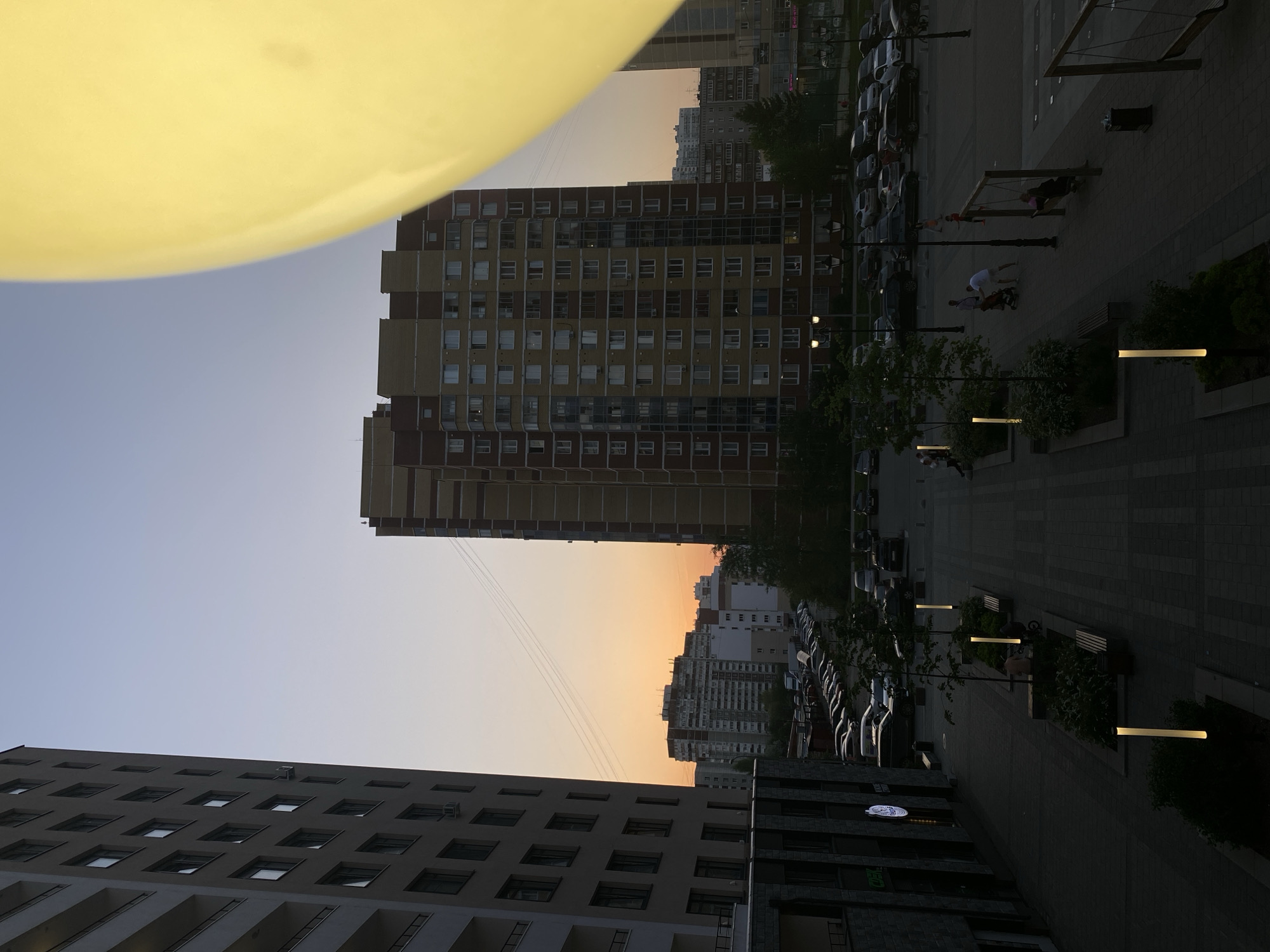 The image shows an outdoor scene in Yekaterinburg, Russia, with multiple buildings of varying heights and architectural styles visible against a backdrop of a pale sky. A portion of a large, yellow object, possibly a balloon or an artificial light source, is present in the upper left corner. The foreground features the tops of two multi-story buildings with numerous windows. In the middle ground, there is a taller building with a mix of brown and yellow tones, also densely populated with windows. Street-level activities are visible on the right side of the image, including parked cars and pedestrians. Some people appear to be engaging in activities such as walking and using playground equipment. Modern street lamps illuminate the pathway.