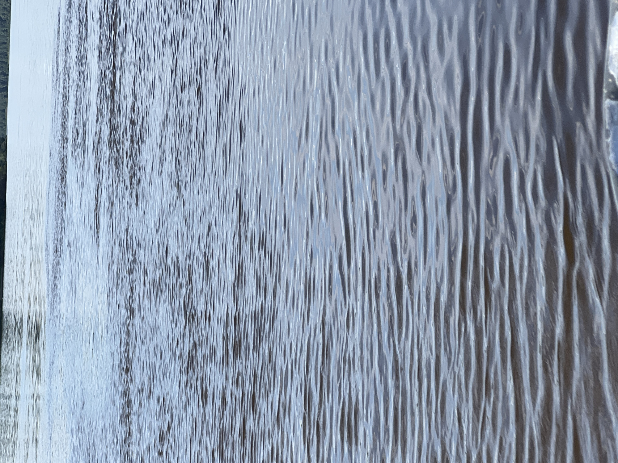 The image displays a close-up, abstract view of what appears to be rippled glass or a similar textured translucent material. The texture is characterized by vertical, undulating lines that create a sense of depth and distortion. Light reflects off the surface, causing variations in brightness and tone, with areas of bright white and darker, shadowy troughs. The overall color palette is predominantly cool, featuring shades of blue, gray, and white, suggesting a bright, possibly overcast day or artificial lighting.
On the left side of the image, there is a hint of a different texture or surface, appearing more solid and lighter in color, possibly a frame or an adjacent object. This contrasts with the fluid, wavy pattern of the main subject. The lighting suggests that the light source is positioned in front of and slightly to the left of the surface, illuminating the raised ridges and casting shadows in the recessed areas.
There are no discernible people, objects, or specific scene elements beyond the textured surface itself. No activity or event is clearly depicted, as the focus is on the abstract pattern and light play. The location context of Sosnovka, Russia, does not provide specific visual cues within this close-up, abstract composition. The image evokes a sense of calmness and perhaps a slightly disorienting or mesmerizing visual experience due to the optical effects of the textured surface. No text is visible in the image.