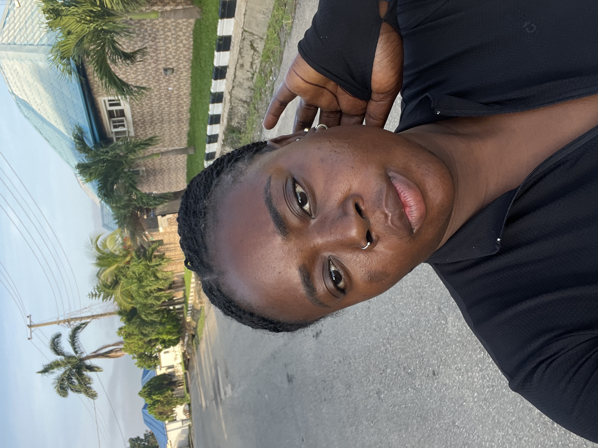This is a portrait of a Black person in an outdoor setting, likely during the daytime. The subject is looking towards the camera with a neutral expression. They have braided hair, a nose ring, and earrings. They are wearing a black long-sleeved shirt with a zipper. The background features palm trees, a building with a blue roof, and a street with a sidewalk that has black and white stripes. Overhead power lines are also visible. The image appears to be taken at eye level, with the subject positioned slightly off-center. There is no visible text in the image.