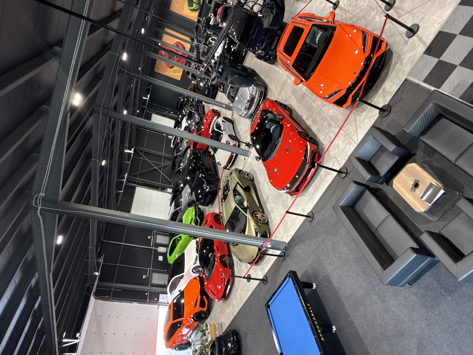 This media file captures an overhead view of a high-end car showroom or private collection located in Plovdiv, Bulgaria. The space is filled with numerous luxury sports cars and exotic vehicles, predominantly arranged on a polished concrete floor.
In the foreground and mid-ground, a vibrant display of performance cars includes an orange Lamborghini Huracan with black accents, a striking red sports car, a gold/tan supercar, a lime-green model, and another bright orange sports car. Many of these vehicles are cordoned off with red ropes attached to black stanchions, indicating a structured display. Further into the showroom, several black cars and a silver Rolls-Royce can be identified.
To the right of the main car display, a contemporary lounge area features dark grey modular sofas and a sleek wooden coffee table, suggesting a comfortable viewing or waiting space. Adjacent to this lounge is a bright blue billiard or pool table, with the text "@PROMOBILE" clearly visible on its side. The flooring in this section includes a dark grey carpet and a distinct black and white checkered pattern near the orange Lamborghini.
The building's interior boasts high ceilings with exposed dark metal beams and trusses, contributing to an industrial-chic aesthetic. Overhead lighting fixtures provide illumination, highlighting the vehicles. The scene conveys a sense of luxury, exclusivity, and a passion for high-performance automobiles. No people are visible in the image.