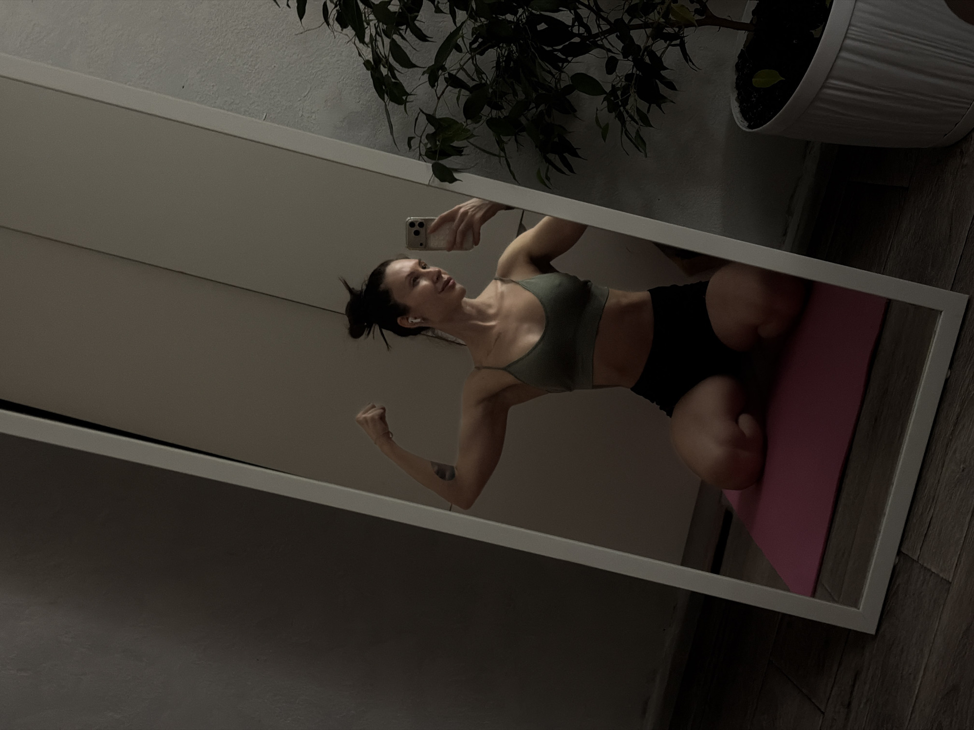 A woman is reflected in a white-framed mirror taking a photo of herself with a smartphone. She is kneeling on a pink exercise mat, wearing a dark olive green sports bra and black shorts. Her hair is pulled back in a messy bun, and she appears to be wearing wireless earbuds. Her left arm is raised in a flexing pose. In the background, out of the mirror's reflection, a potted plant is visible. The setting appears to be an indoor residential space. The image was taken in Perm, Russia.