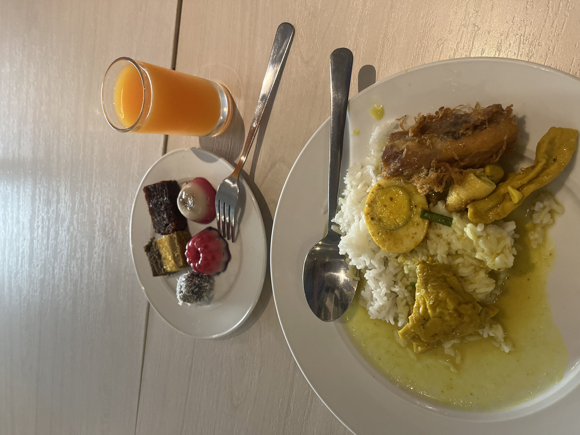The image displays two white plates of food and a glass of orange juice on a light-colored wooden surface, likely a table. The foreground plate holds a serving of white rice topped with a halved hard-boiled egg, pieces of yellow curry chicken, and a piece of fried food. A silver spoon rests on the edge of this plate. The background plate contains an assortment of small desserts: a rectangular layered cake, a small dark brown cake, a round red dessert with a dented top, a spherical dessert coated in shredded coconut, and a white dessert with a pink bottom. A fork rests between the two plates. The overall scene suggests a meal in a casual dining setting. No individuals are visible in the image. The lighting indicates an indoor environment with overhead illumination, casting subtle shadows. The provided crops highlight specific elements of the food and cutlery on the table. There is no information within the image or its crops that can be used to determine the specific location in the city of Gapurosukolilo, Indonesia.