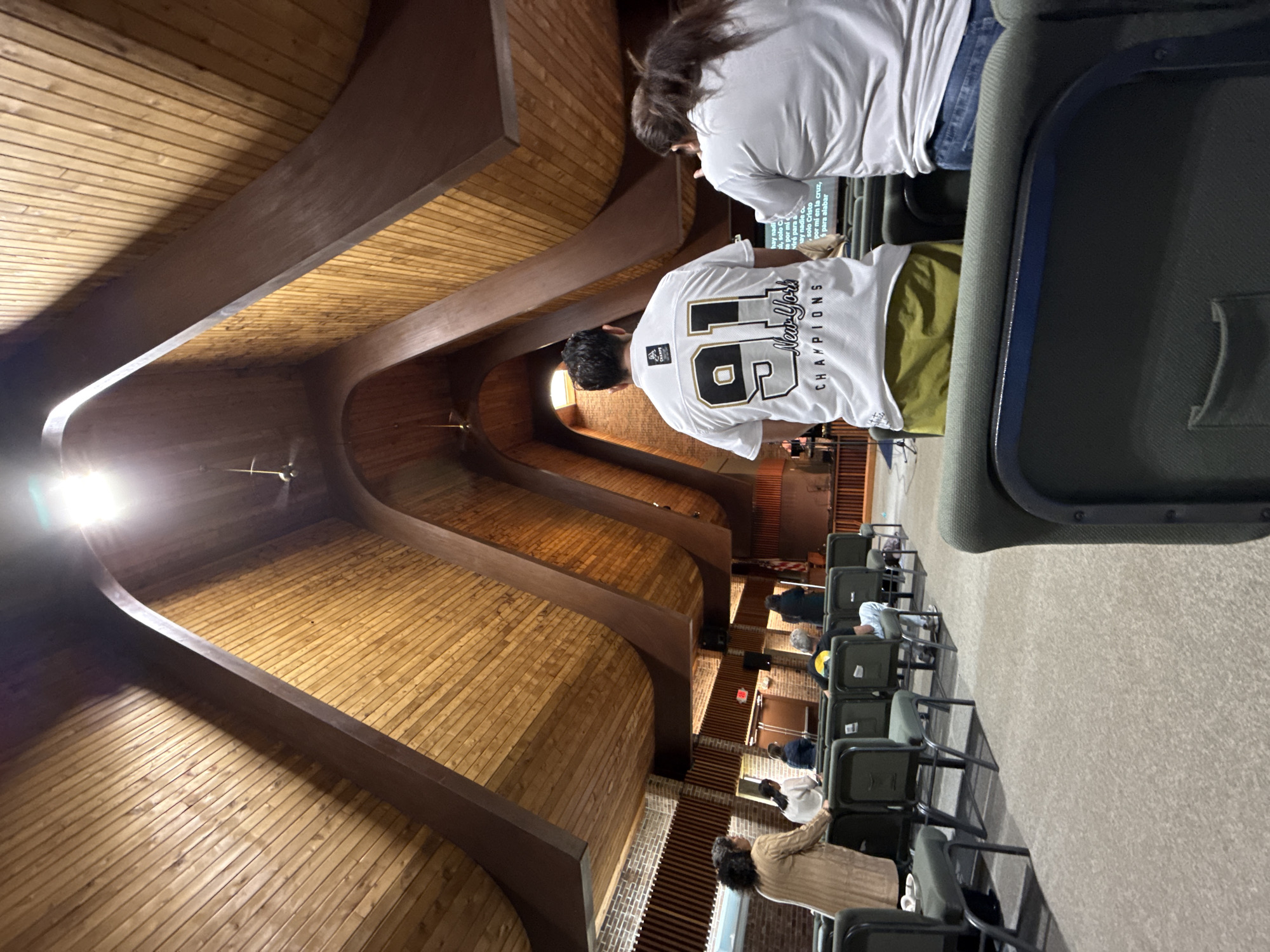 The image depicts the interior of a church, possibly located in Columbus, Ohio, United States. The architecture features a high, curved wooden ceiling with exposed beams. Rows of chairs are set up for seating, and several individuals are present, either sitting or standing. At least two people are wearing white t-shirts with writing or designs on the back. One shirt has "91" and "New York CHAMPIONS" written on it. A screen or monitor is visible in the background, displaying text in Spanish. The scene appears to be a church service or event.