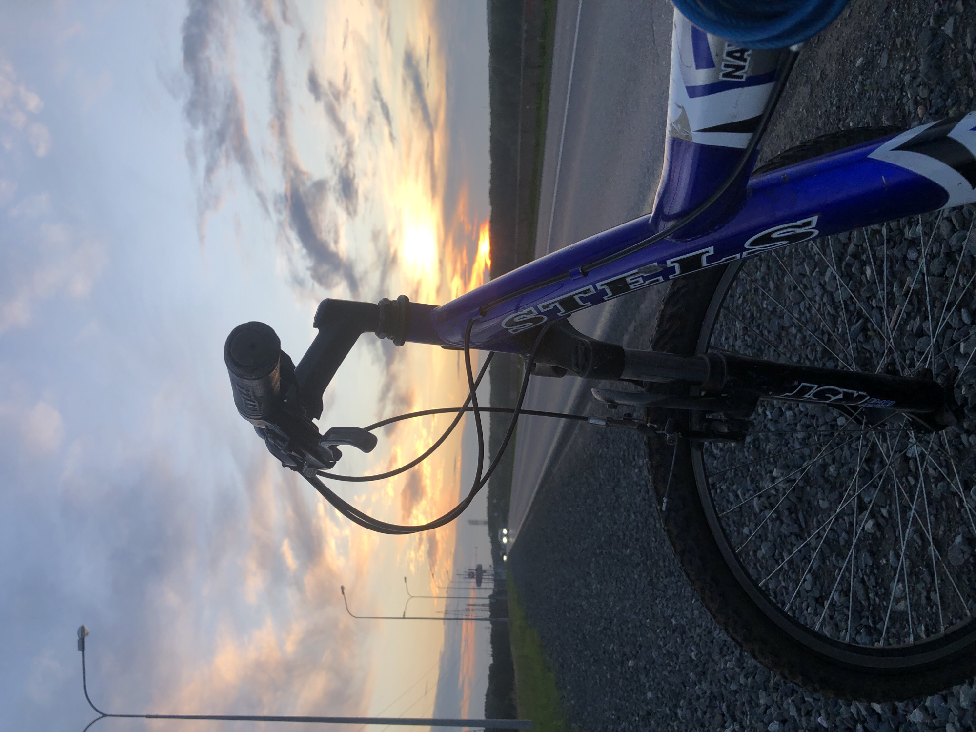 A bicycle is parked on a gravel shoulder next to a paved road. The bicycle is blue and white with a visible "GT" logo. The handlebars and front wheel are in the foreground. The road stretches into the distance, with two cars visible. In the background, there are power lines, trees, and a radio tower. The sky is filled with clouds, illuminated by the setting sun, casting an orange and pink glow. The location is near the city of Chaikovsky, Russia, along a road leading away from it.