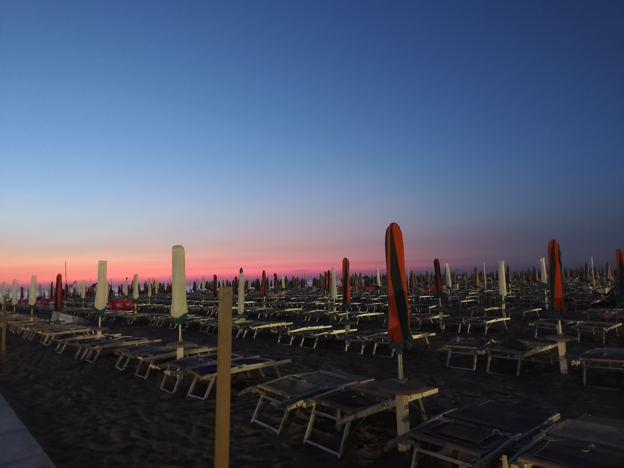 The image depicts a beach in Rimini, Italy, at dusk. The sky displays a gradient of colors, transitioning from deep blue at the top to hues of pink and orange near the horizon. Rows of empty lounge chairs and closed umbrellas are neatly arranged on the dark sand. The scene is devoid of people, suggesting the beach is either closed for the evening or in a state of transition between day and night. The atmosphere is serene and quiet.