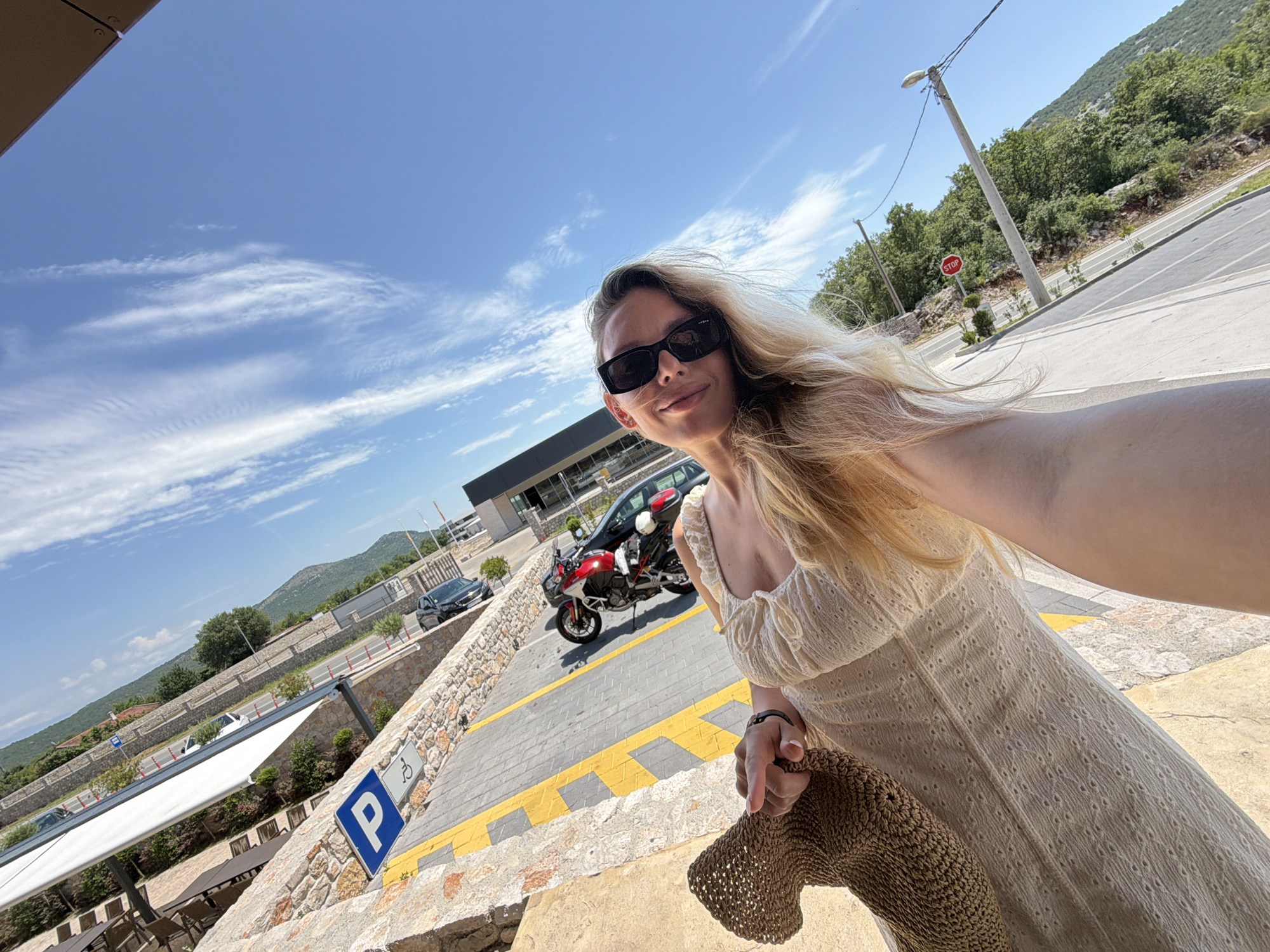 The image depicts a woman holding a woven hat and taking a selfie outdoors, with a picturesque background. She is wearing a light-colored, ruffled dress and sunglasses, her long blonde hair framing her face. In the background, a parking spot with a blue accessibility symbol is visible, along with a street and parked motorcycles. The scene is set against a backdrop of lush green hills under a bright blue sky with wispy clouds. The overall impression is one of a sunny day, possibly in a tourist destination like Bigor, Montenegro, given the terrain and clear weather.