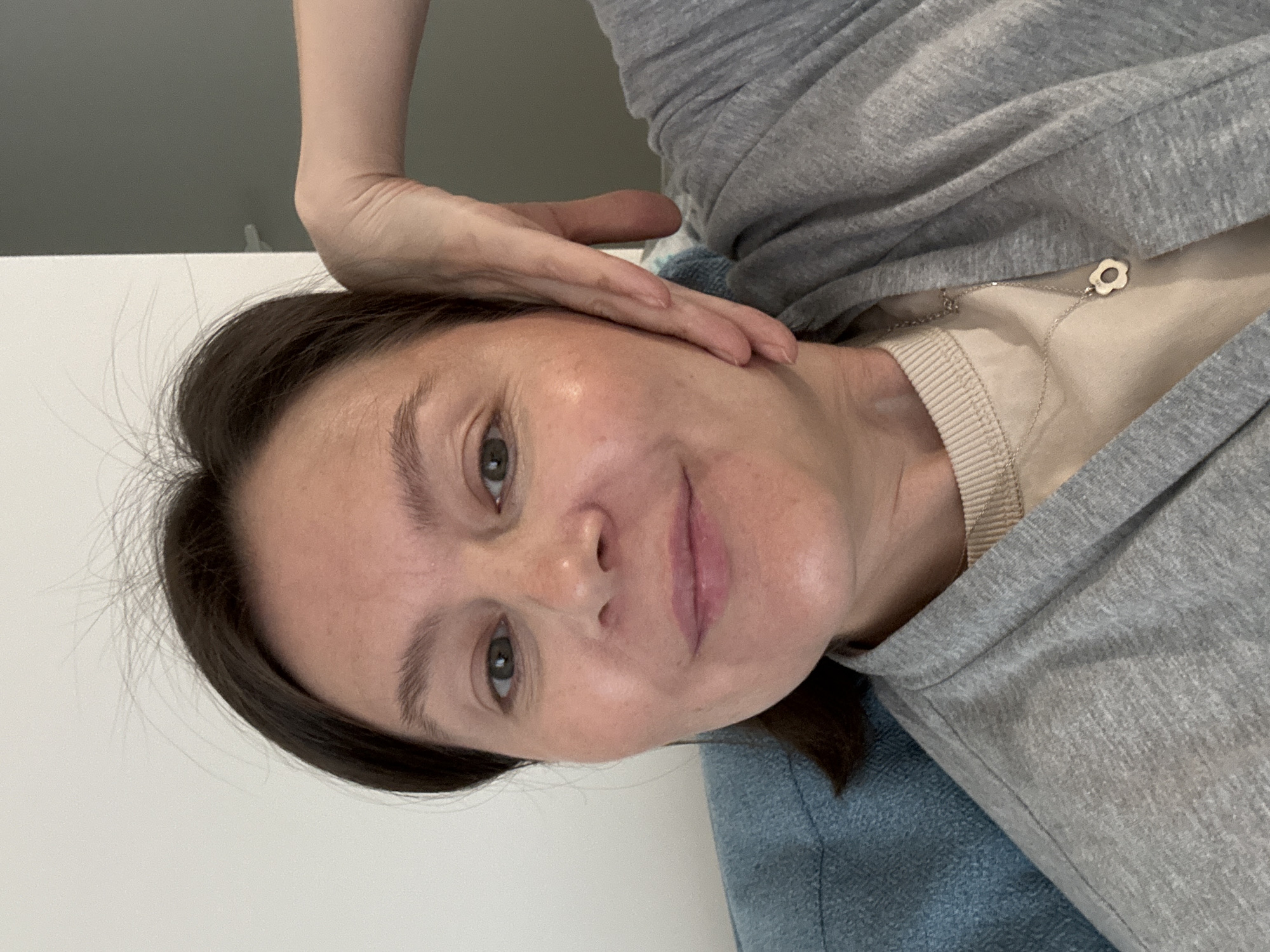 A woman is pictured close-up, likely taking a selfie. She has fair skin, brown hair pulled back, and is wearing a gray hooded sweatshirt over a tan top. A silver necklace with a small flower pendant is visible. Her hand is resting on her head near her ear. The background is plain and white. There is no indication of the location being Izhevsk, Russia, or any other specific city.