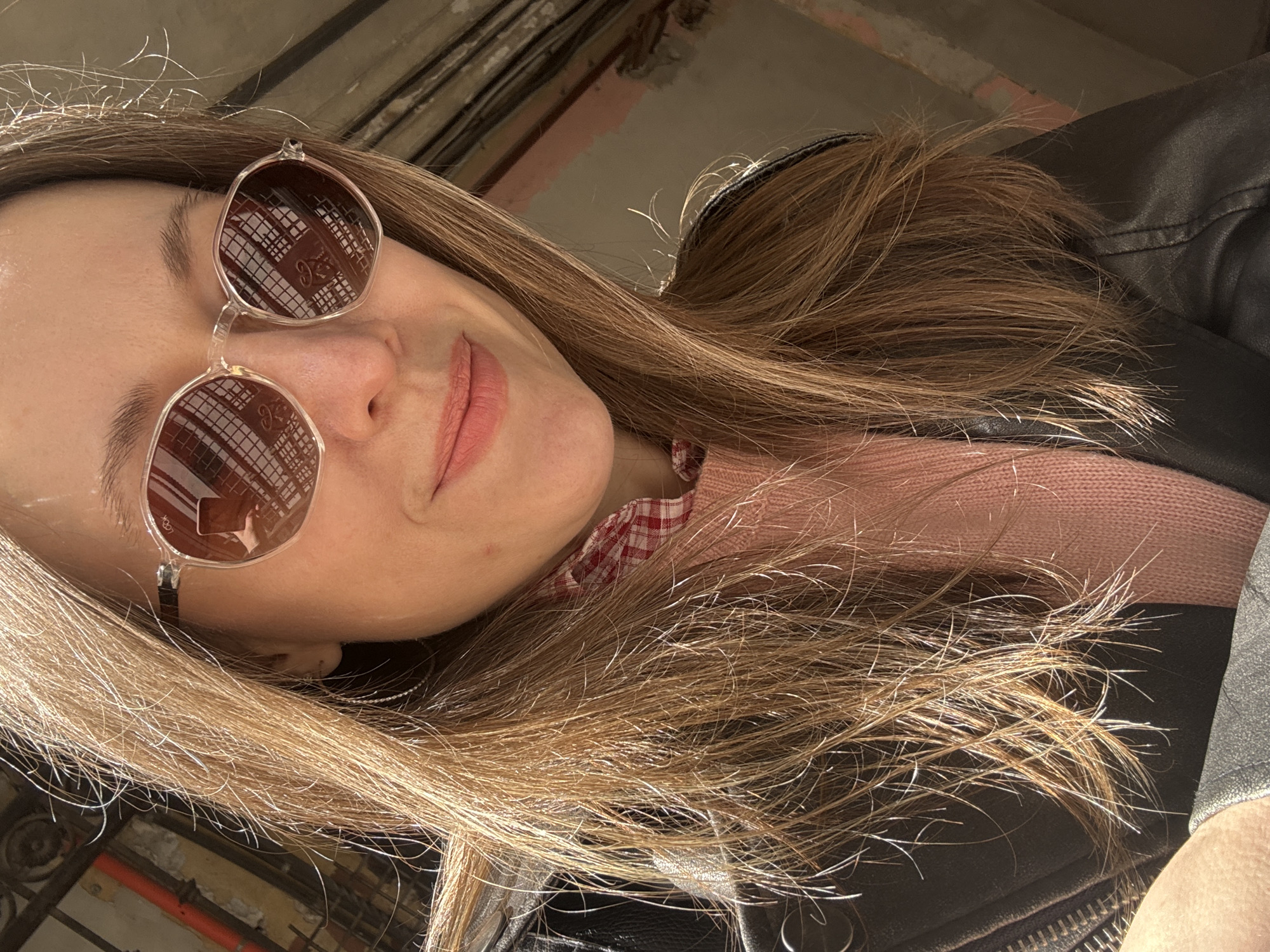 A woman is pictured from a low angle, wearing sunglasses, a pink sweater with a red and white checkered collar, and a black leather jacket. Her hair is long and light brown. The reflection in her sunglasses reveals a building with a grid-like pattern, indicative of a windowed structure. The background appears to be a partially renovated space with exposed pipes and brickwork. The location can be inferred to be somewhere in Saint Petersburg, Russia, based on the architectural style of the reflected building and general urban aesthetic.