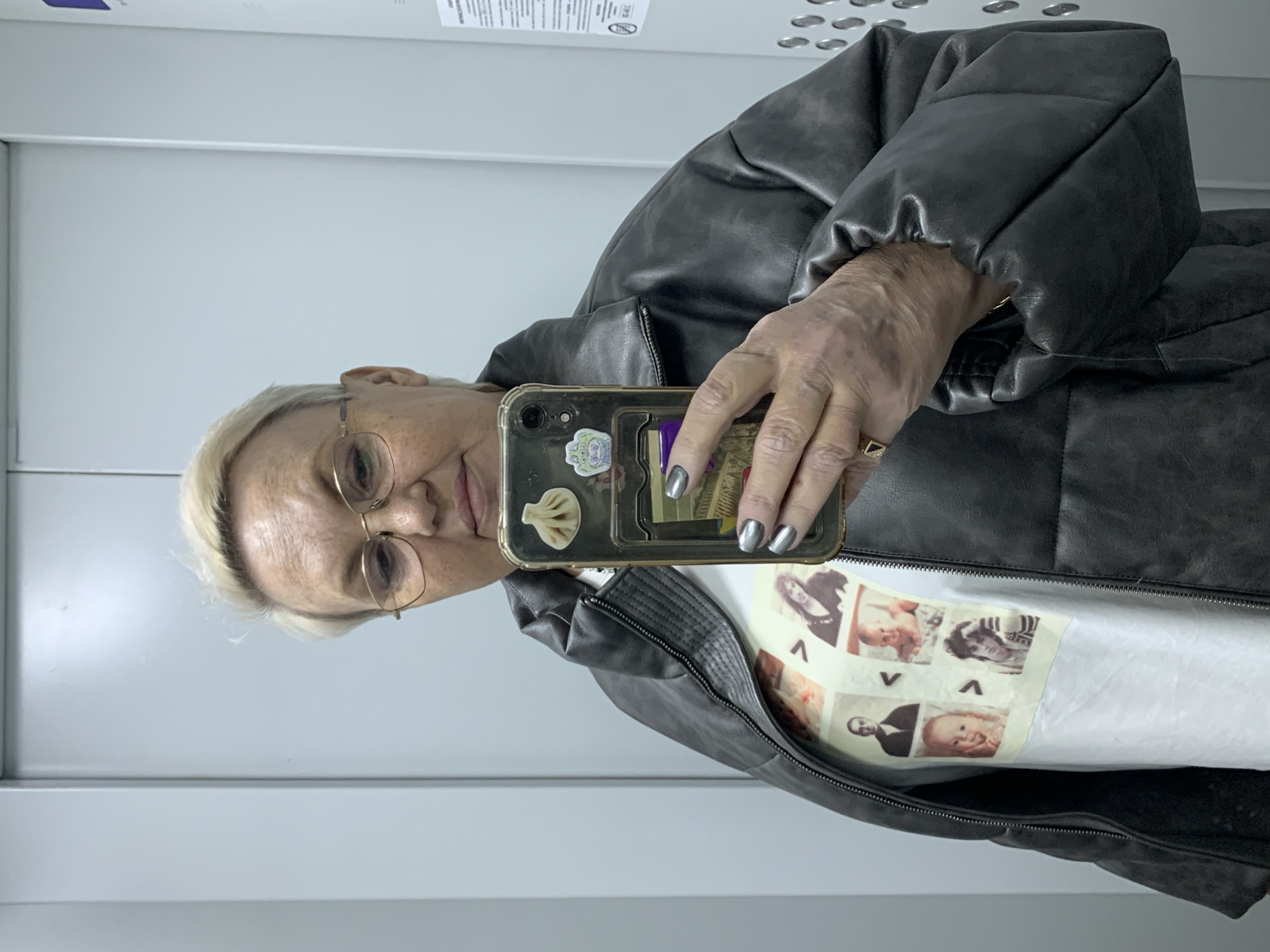 A person is taking a selfie in what appears to be an elevator with white walls. The individual has fair skin, short blonde hair, and wears gold-rimmed glasses. They are wearing a white shirt with a collage of black and white images, covered by a dark grey or black, possibly leather, puffer-style jacket. The person holds a smartphone in their right hand, with silver nail polish on their fingernails. The phone case has stickers attached. The location is unspecified except that it is in the city of Chaikovsky, Russia.