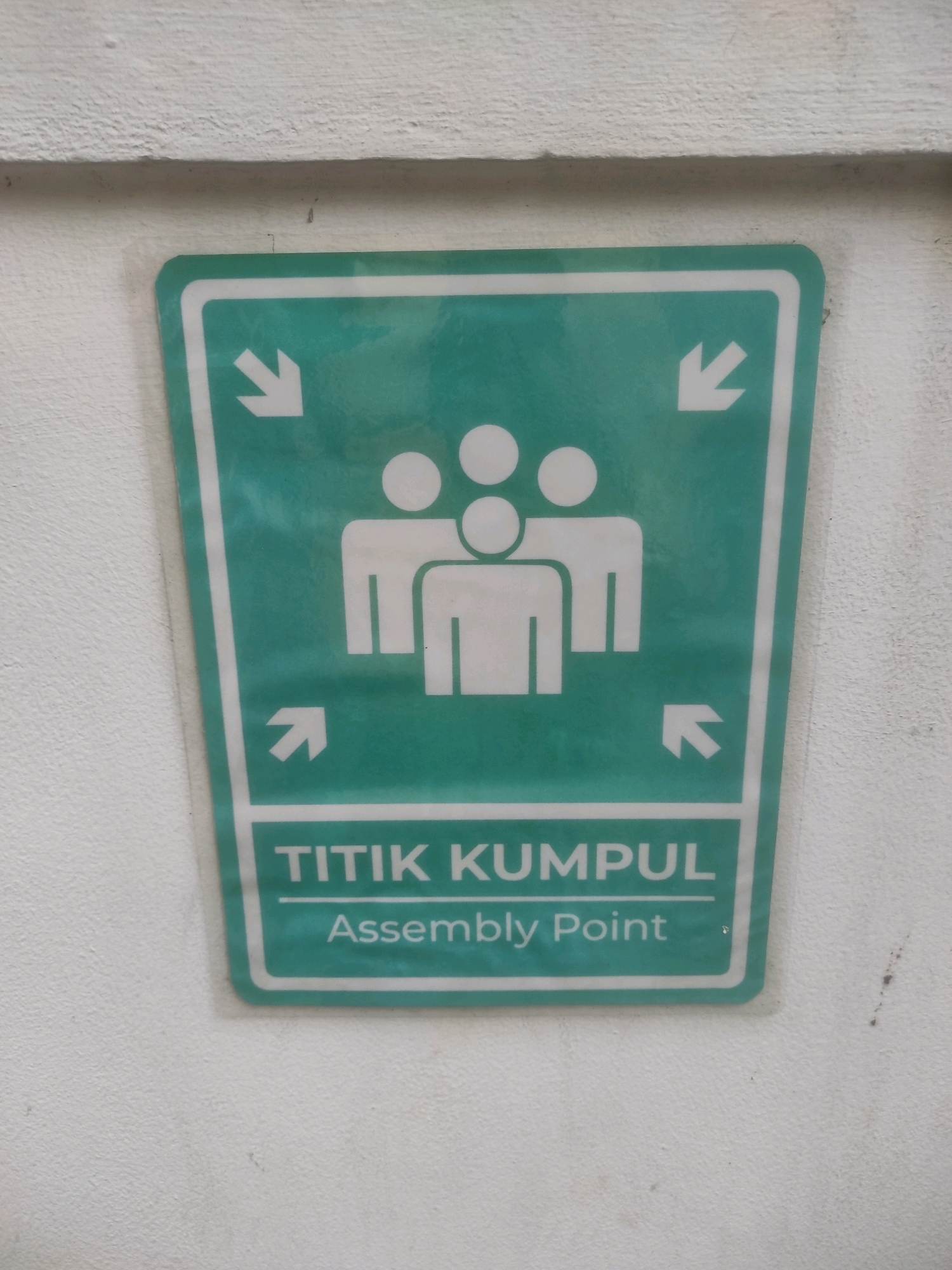 A green and white sign for an assembly point is affixed to a textured white wall. The sign is shaped like a rounded-off trapezoid and has a thick white border. Inside the border, four white arrows point inwards towards the center of the sign. In the center of the sign is a white pictogram of three figures, representing people, clustered together. Below the pictogram, the words "TITIK KUMPUL" are written in white block letters, followed by "Assembly Point" in white lowercase letters. The sign appears to be slightly reflective. The wall itself is rough and shows some minor staining.