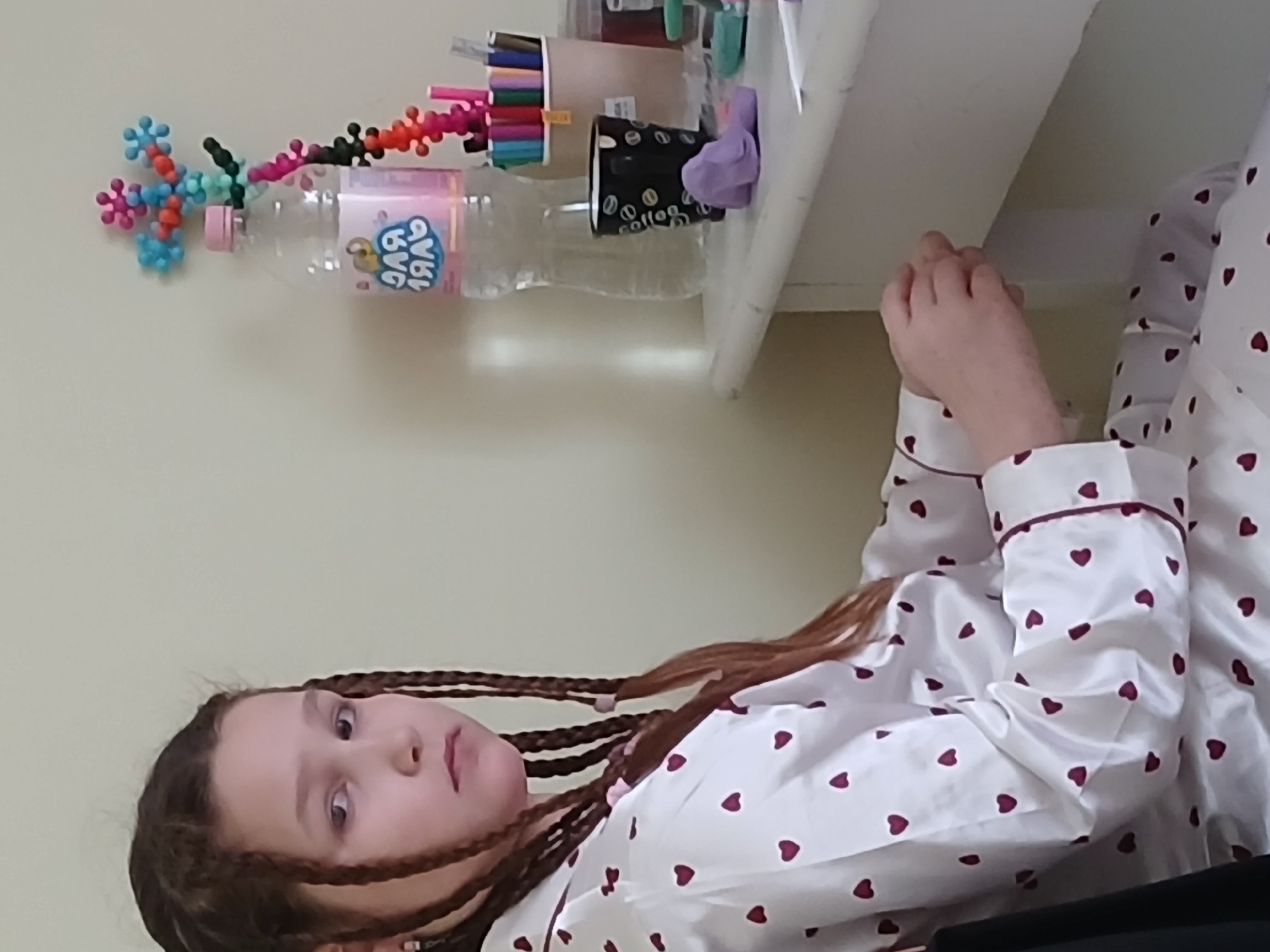 A young girl with long, dark brown hair styled in numerous thin braids is captured in an indoor setting, likely a bedroom or personal space. She is seen from a slightly elevated perspective, lying down or reclining, and looking directly towards the camera with a neutral expression. She wears white pajamas patterned with small, dark red hearts and accented with maroon piping along the collar and cuffs. Her hands are clasped together, resting near her chest.
Above her, positioned on a white shelf or desk, are several objects. A clear plastic water bottle with a pink cap is visible, featuring a pink label with stylized white text that appears to read "BAG BAG." Attached to the bottle is a colorful, elaborate construction made of interconnected plastic pieces, resembling spiky spheres or pop beads, in shades of blue, light blue, pink, red, black, and orange. Beside the bottle, a light brown cylindrical container holds a variety of colored markers or pens. A black paper cup, adorned with white script spelling "coffee" and illustrations of coffee beans, sits next to a purple scrunchie or small piece of fabric. The background consists of a plain, light-colored wall. The scene conveys a sense of quiet relaxation within a personal and creative environment.