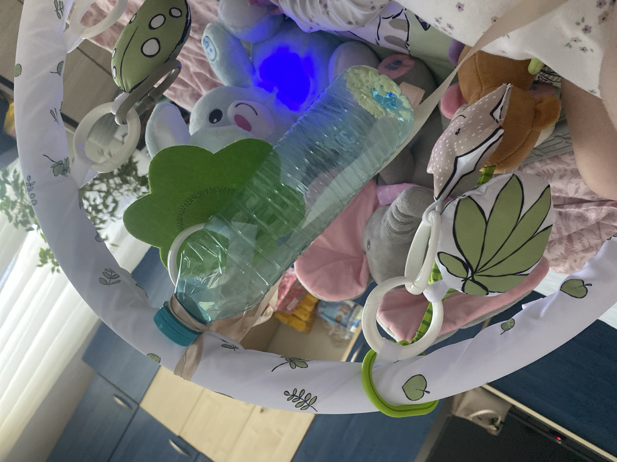 The image presents an overhead perspective of a baby's play gym, with a baby partially visible underneath. The central feature is a white fabric-covered arch, decorated with green leaf motifs, from which several toys are suspended.
Starting from the left, a green caterpillar-shaped soft toy with white spots hangs from a white plastic ring. Adjacent to it, a green fabric toy shaped like a flower is visible. A transparent plastic bottle, seemingly repurposed, hangs near a light blue stuffed animal, which emits a prominent blue glow from its body, suggesting an internal light source. Further to the right, a brown stuffed animal with a distinct light-colored, patterned face, possibly a deer or fox, is suspended. Another green leaf-shaped fabric toy is also part of the collection.
A baby's leg and a portion of its body, clad in a patterned light-colored garment, are visible beneath the play gym, indicating the baby is lying down and potentially interacting with the toys.
The background reveals an indoor setting, likely a nursery or play area. Elements include light-colored curtains on the left, dark blue cabinets or furniture, and what appears to be a wooden floor or surface. In the lower right corner, a black electronic device with a red indicator light is visible. The scene is illuminated by ambient indoor lighting, augmented by the glowing blue toy. The location context for this domestic scene is Blagoevgrad, Bulgaria. No discernible text is visible in the image.