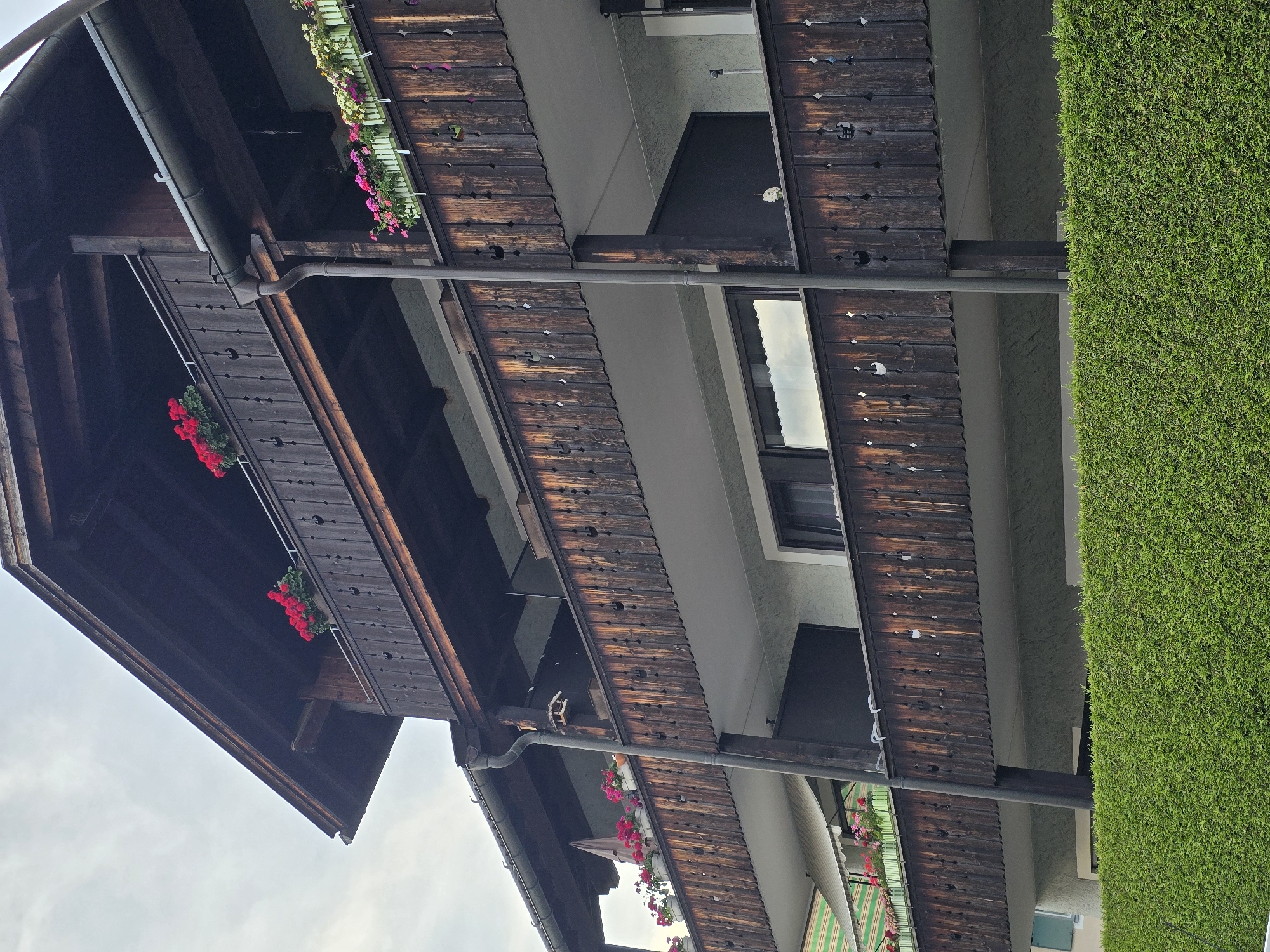 This media file captures an exterior view of a multi-story building with a rustic wooden facade, featuring flower boxes overflowing with vibrant red and pink blossoms. The building appears to be a guesthouse or residential property, possibly in the Alps given the architectural style and the presence of a lush green hedge. The scene is set during the day, under an overcast sky.
The wooden structure is characterized by its weathered planks and decorative cutouts. Balconies are visible on different levels, adorned with hanging baskets of flowers, suggesting a well-maintained and welcoming aesthetic. A downspout system is integrated along the roofline. Windows on the upper floors reflect the cloudy sky. To the right of the building, a thick, neatly trimmed green hedge forms a striking contrast to the dark wood.
The image is oriented such that the sky is at the bottom and the ground level is at the top, indicating it was taken looking upwards. No people are visible in the frame, and there is no visible text. The overall impression is one of charming, traditional alpine architecture set against a backdrop of natural greenery. The blooming flowers add a cheerful and colorful element to the scene. The overcast weather suggests a mild or possibly damp day. The location cues like the style of the building and the hedge point towards a European setting, specifically Garmisch-Partenkirchen, Germany.