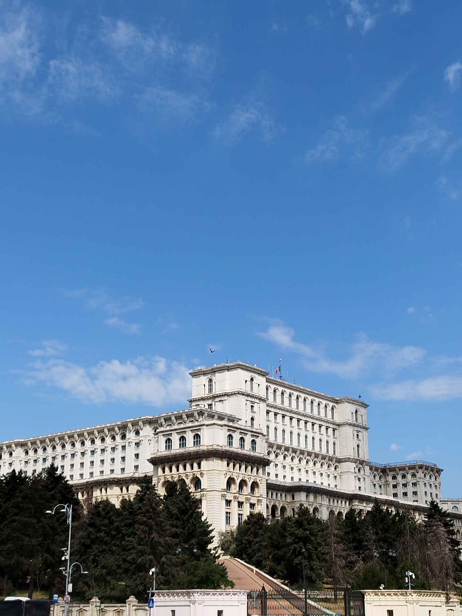 The image shows the Palace of the Parliament in Bucharest, Romania, under a clear blue sky with scattered white clouds. The massive, ornate building dominates the lower half of the frame, displaying its neoclassical architecture with numerous windows, arched details, and multiple levels. Dark green coniferous trees are situated in front of the building, partially obscuring the lower facade. In the foreground, there are streetlights, a traffic signal, and parts of a fence or barrier. The overall scene depicts a bright, daytime view of this iconic landmark.