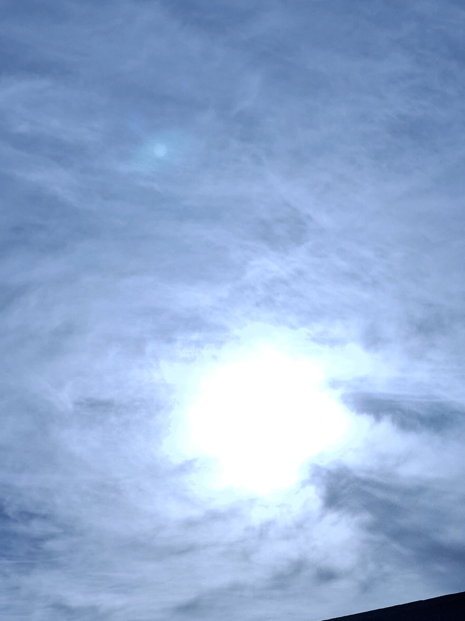 The image captures a sky filled with wispy clouds, predominantly in shades of blue and white. The sun is a bright, glowing orb near the center of the frame, partially obscured by clouds, creating a lens flare effect. A dark, angled shape, likely the edge of a building or structure, is visible in the lower right corner, suggesting an urban environment. The scene is devoid of any people or specific landmarks that would pinpoint its exact location within Torrevieja, Spain. The lighting indicates it is daytime, possibly late morning or early afternoon, with the sun at a high angle.