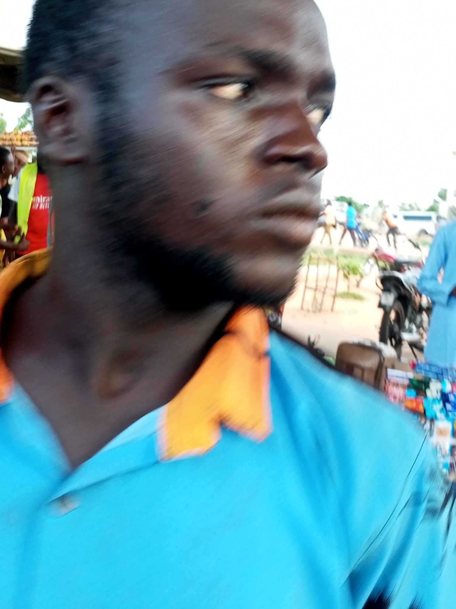 The image shows a close-up, slightly blurry, portrait of a dark-skinned man with a beard, looking to his right. He is wearing a bright blue shirt with an orange collar. In the background, out of focus, there are other people, some motorcycles, and what appears to be a market stall with colorful items. The setting suggests an outdoor urban environment, possibly a busy street or market area in Gombe, Nigeria, given the context of the request. The lighting suggests it is daytime.