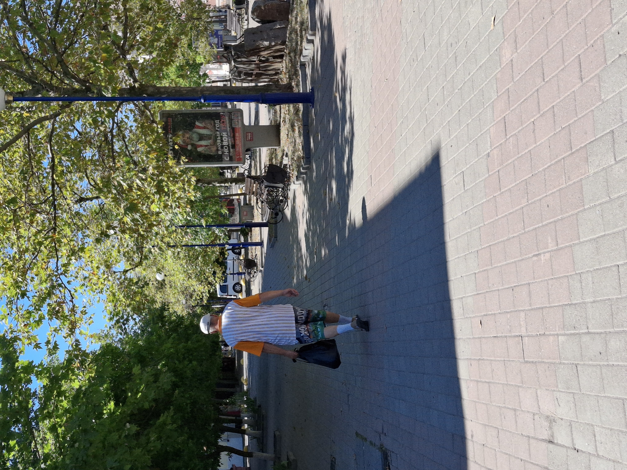 An outdoor, eye-level, medium shot captures a man walking away from the camera on a paved sidewalk lined with trees. The man is wearing a white cap, a horizontally striped shirt in white and orange, and patterned shorts. He carries a black plastic bag in his right hand. The sidewalk is paved with light-colored rectangular bricks, and a large shadow from overhead trees falls across it. To the right of the man, a blue lamppost stands, and behind it, a billboard displays an advertisement for the New Year with an image of Santa Claus. Further back, trees with green foliage form a canopy, and the sky is a clear blue. In the background, other elements of an urban environment are visible, including benches, a white van, and more trees. The lighting suggests it is daytime, with bright sunlight casting shadows.