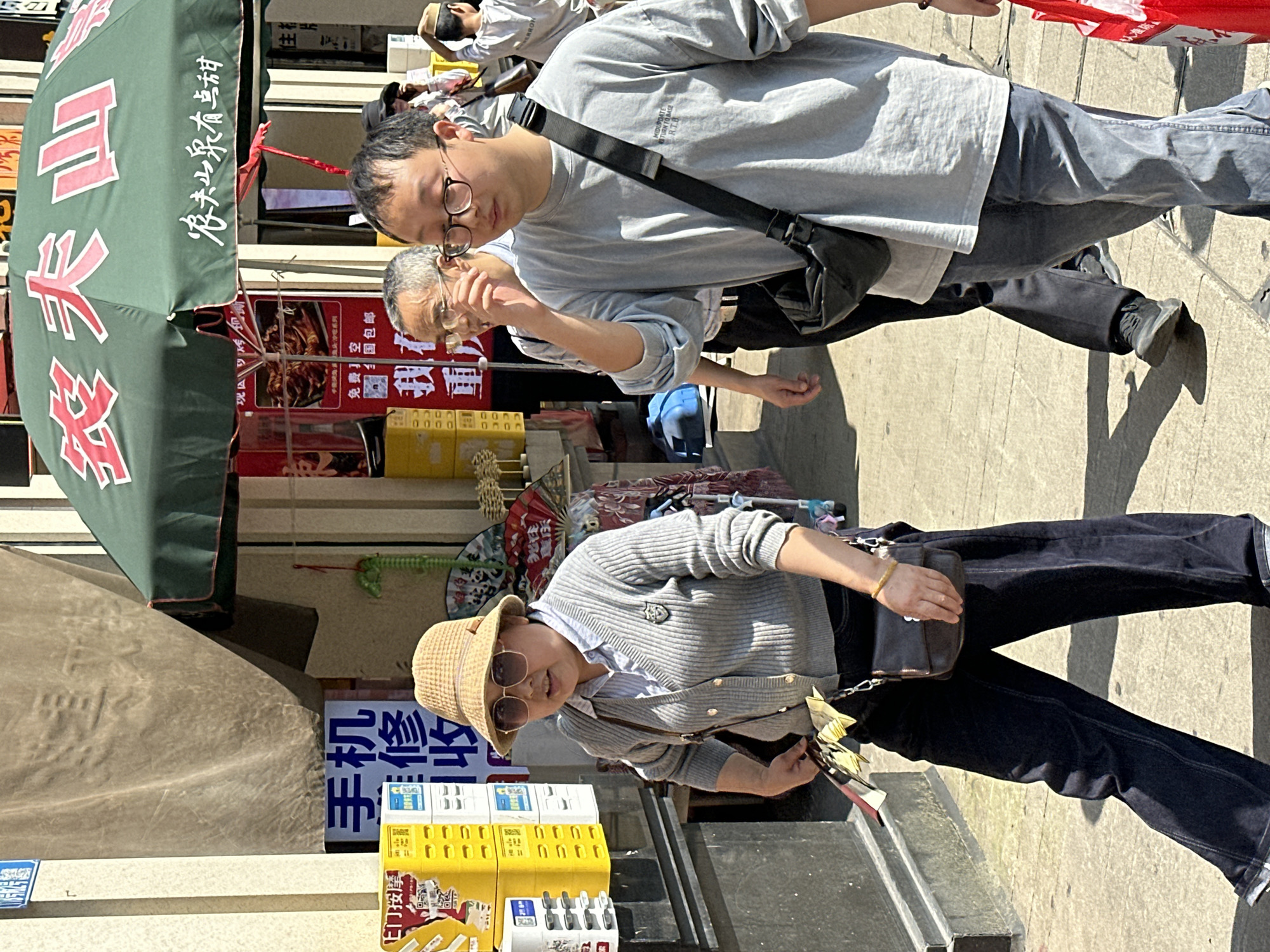 This image, likely captured in the city of Yuzhong District, China, shows two people walking on a city street. A younger man, wearing glasses and a gray shirt with a black cross-body bag, is seen walking next to an older man, also wearing a gray shirt and glasses. Next to them stands a woman wearing a straw hat, sunglasses, a gray cardigan, and dark pants. She is holding a small bag. Behind them, there are several signs with Chinese writing, an umbrella with writing, and several boxes.