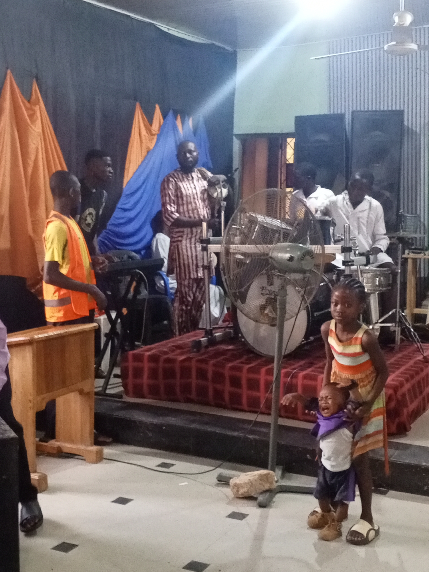 The image depicts a church interior in Unknown, Unknown. A band is set up on a raised platform covered with red patterned fabric. The band consists of five men; one wearing a striped brown and white robe stands at the front of the platform, another man wearing a yellow shirt and orange safety vest stands to the left, a third plays a keyboard, and two men in white shirts are playing drums. Behind the band are two large black speakers and a wall with vertical stripes. In the foreground, a young girl in a striped dress is holding a crying toddler. An industrial fan stands between the girl and the band. The floor is tiled with white and black squares. Orange and blue drapes hang behind the band. A ceiling fan is visible in the upper right corner.