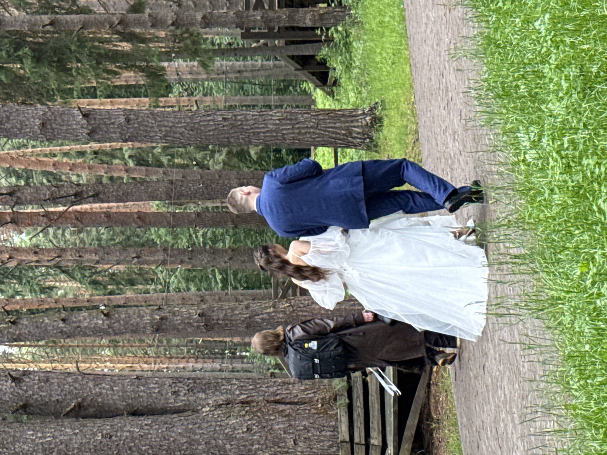The image depicts three individuals walking along a light-colored paved path within a dense forested area, specifically a park or natural preserve in Votkinsk, Russia. All individuals are viewed from behind, progressing away from the camera.
The central figure is a female wearing a white, off-the-shoulder wedding gown with a flowing skirt. Her dark, styled hair is partially visible. To her immediate right is a male figure, dressed in a dark blue suit jacket, matching trousers, and dark shoes. He has short hair. To the left and slightly behind the female in the wedding gown is a third individual, wearing a long, dark brown coat over dark trousers and dark shoes. This individual carries a black backpack, which has a small yellow and white object attached to its left side. The individual has long, brown hair and appears to be holding a white strap or piece of fabric, possibly from the wedding gown, in their left hand.
The environment is characterized by numerous tall, slender evergreen trees with grey-brown, textured bark, forming a dense background. The ground adjacent to the path is covered with green grass and various plants. In the mid-ground, among the trees, rudimentary wooden structures, resembling parts of a platform or recreational installation, are visible. Thin, horizontal wires or strings are stretched between some of the tree trunks. The overall lighting is soft and diffused.