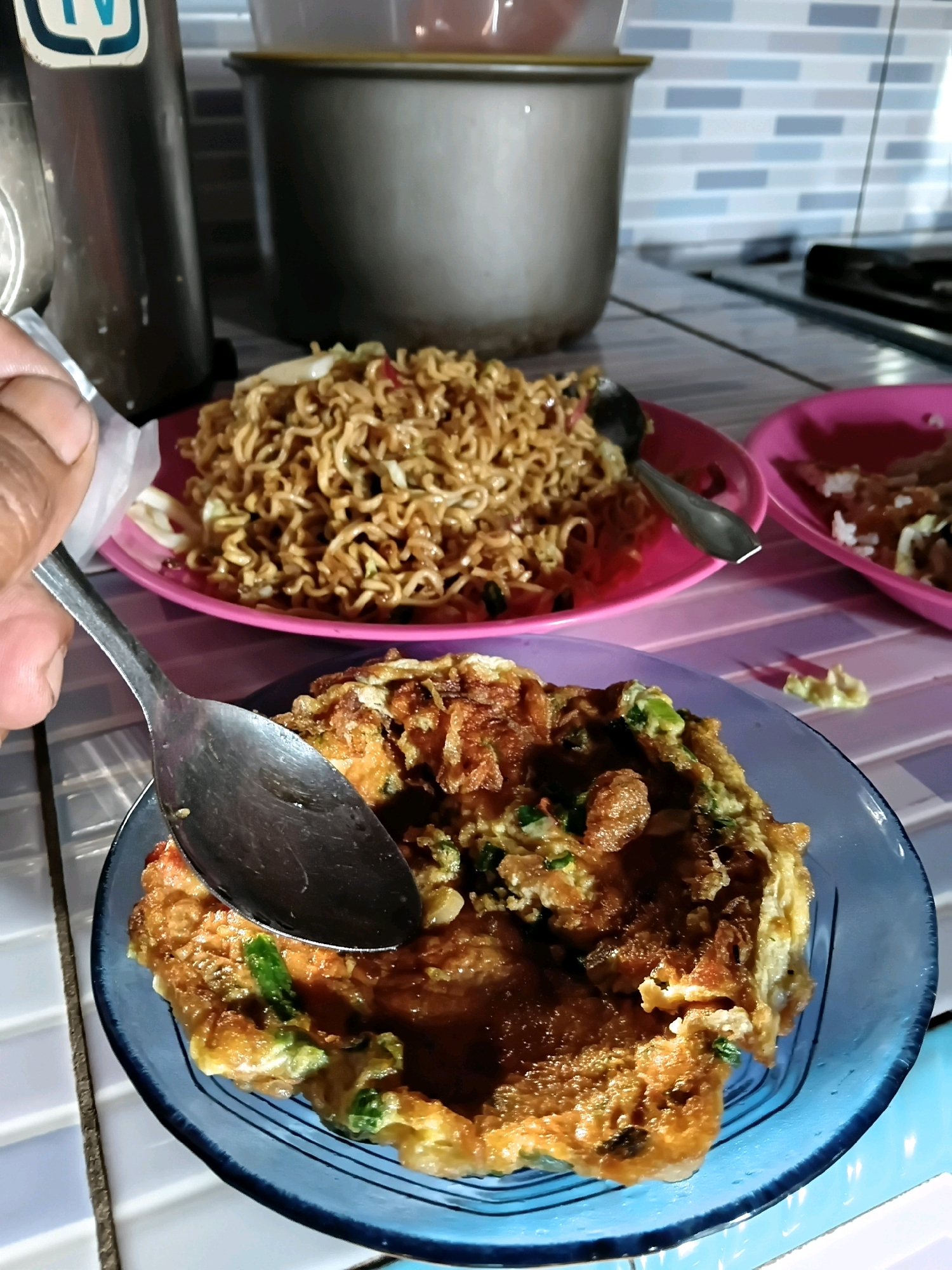 A hand holds a spoon over a blue plate containing an omelet with green onions. In the background, a pink plate holds instant noodles, and another pink plate has rice and what appears to be meat. The scene is set on a table with a tiled countertop and backsplash. The lighting suggests an indoor setting.
