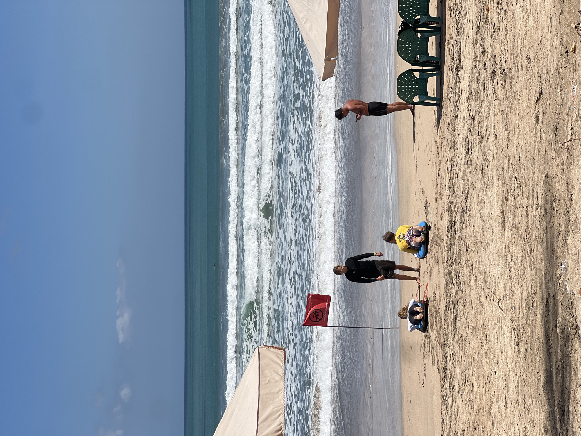 In Kuta, Indonesia, a serene beach scene unfolds under a clear blue sky. The ocean, a vibrant turquoise, presents a series of gentle waves crashing onto the shore. A red flag, indicating "Swimming Prohibited," stands planted on the wet sand, accompanied by a person in a black wetsuit, likely a lifeguard. Nearby, three individuals are positioned on the beach: two lie on boogie boards facing the ocean, and another stands engrossed in their mobile phone. Two large, beige beach umbrellas offer shade, one on the left and another on the right, while three green plastic chairs sit vacant on the sand, facing the water. The sand itself is a light brown, marked by footprints and scattered debris. In the distance, a lone surfer rides a wave.