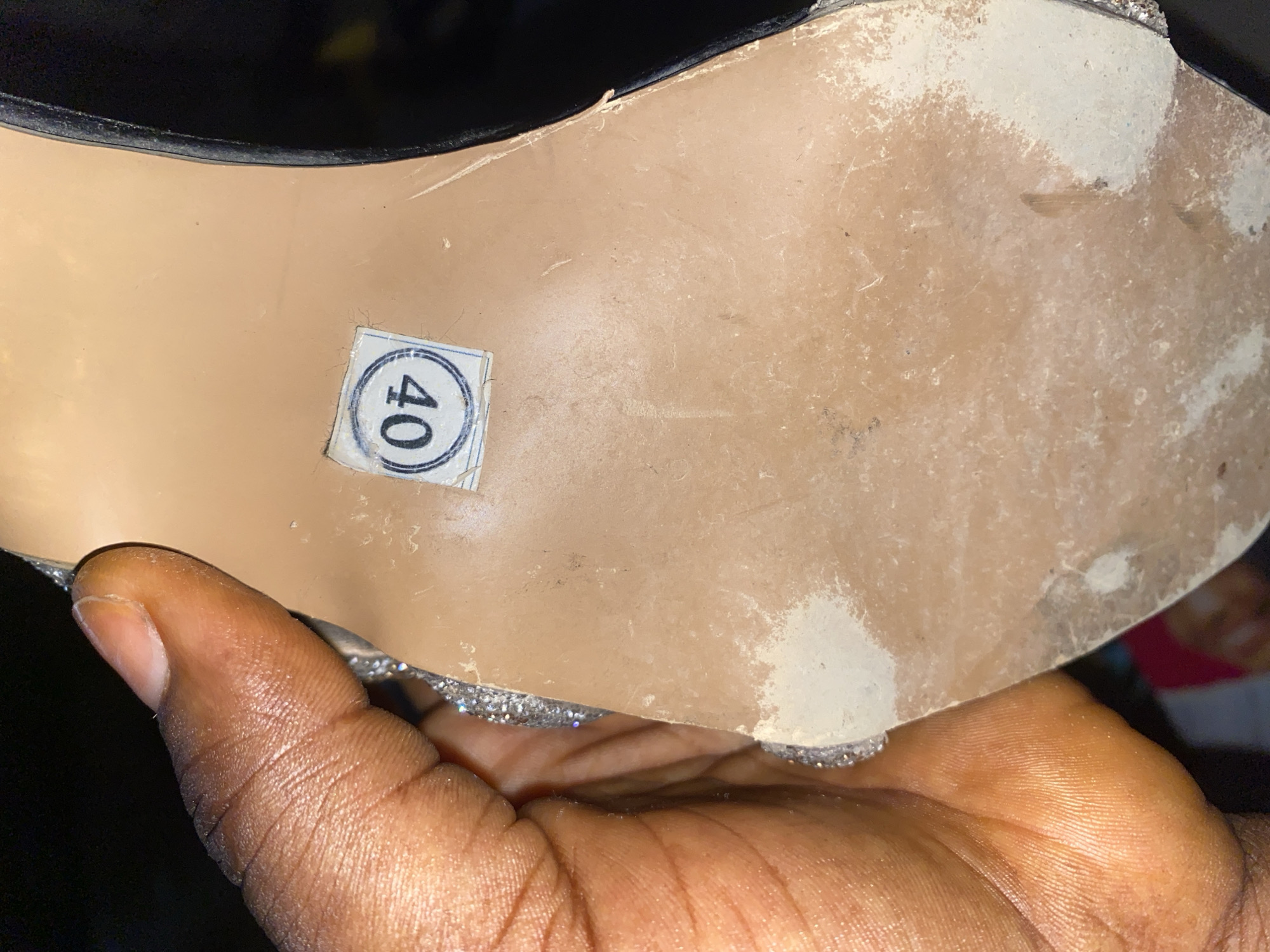 The image displays the sole of a shoe, held by a person's hand. A sticker with the number "40" printed inside a circular emblem is affixed to the sole. The sole appears to be made of a light-colored material and shows signs of wear and tear, with scuff marks and dust. The shoe itself has a silver, glittery trim along its edge. The background is dark, suggesting an indoor or nighttime setting. There is no information in the image to determine the location in Jauro Yinu, Nigeria.