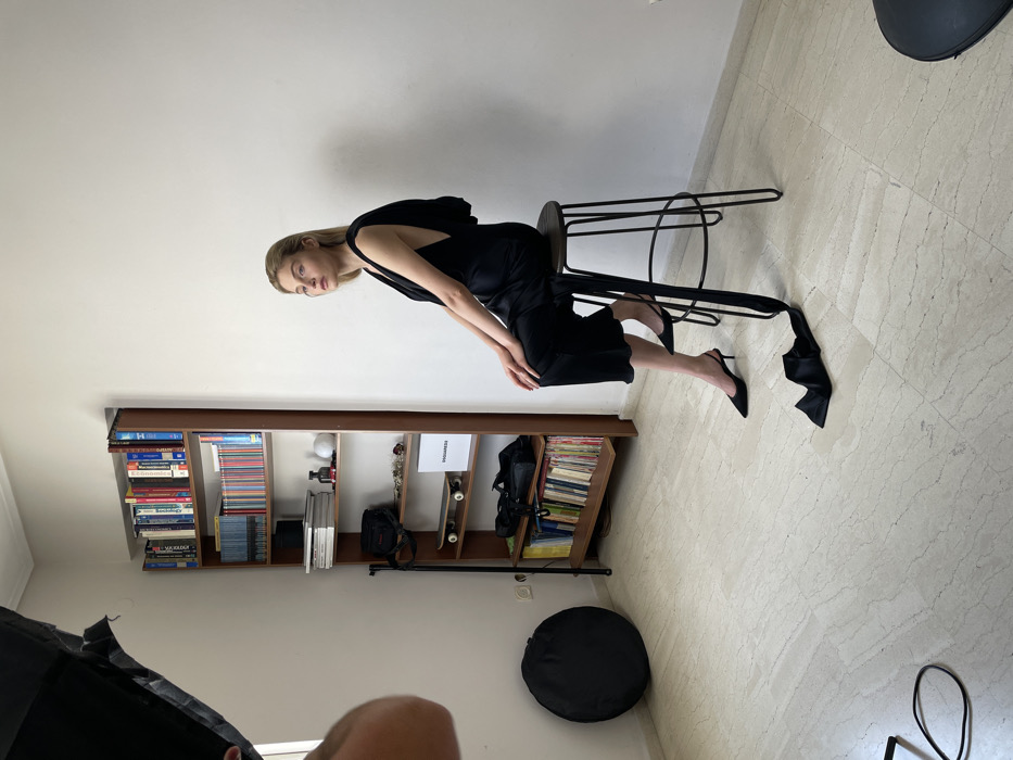 The original image is rotated 90 degrees clockwise. When viewed correctly, it depicts an indoor, full-shot photograph of a young woman posing for a photoshoot, likely in a studio or a well-lit room in Vironas, Greece. The woman is positioned slightly off-center to the right, seated on a tall, black metal stool with a dark wooden top.
The woman has long, straight blonde hair and is dressed in a sleek black sleeveless garment that features a draped or flowing fabric detail from the back, extending towards the floor. She wears black pointed-toe high-heeled mules. She leans forward on the stool, resting her right hand on her left knee, and looks directly towards the camera with a neutral, composed expression. Her left leg is subtly extended, with the black fabric detail from her garment trailing near her foot.
The background features plain, light-colored walls and a light-tiled floor with a subtle pattern. To the left, a wooden bookshelf is visible, well-stocked with numerous books, some small decorative items, and a prominent white box labeled "PROGRESS." On the floor to the lower left, a black circular photography softbox or reflector is present, alongside part of another piece of photography equipment in the bottom-left corner, suggesting a professional photography setup. The lighting appears to be controlled studio lighting rather than natural daylight.