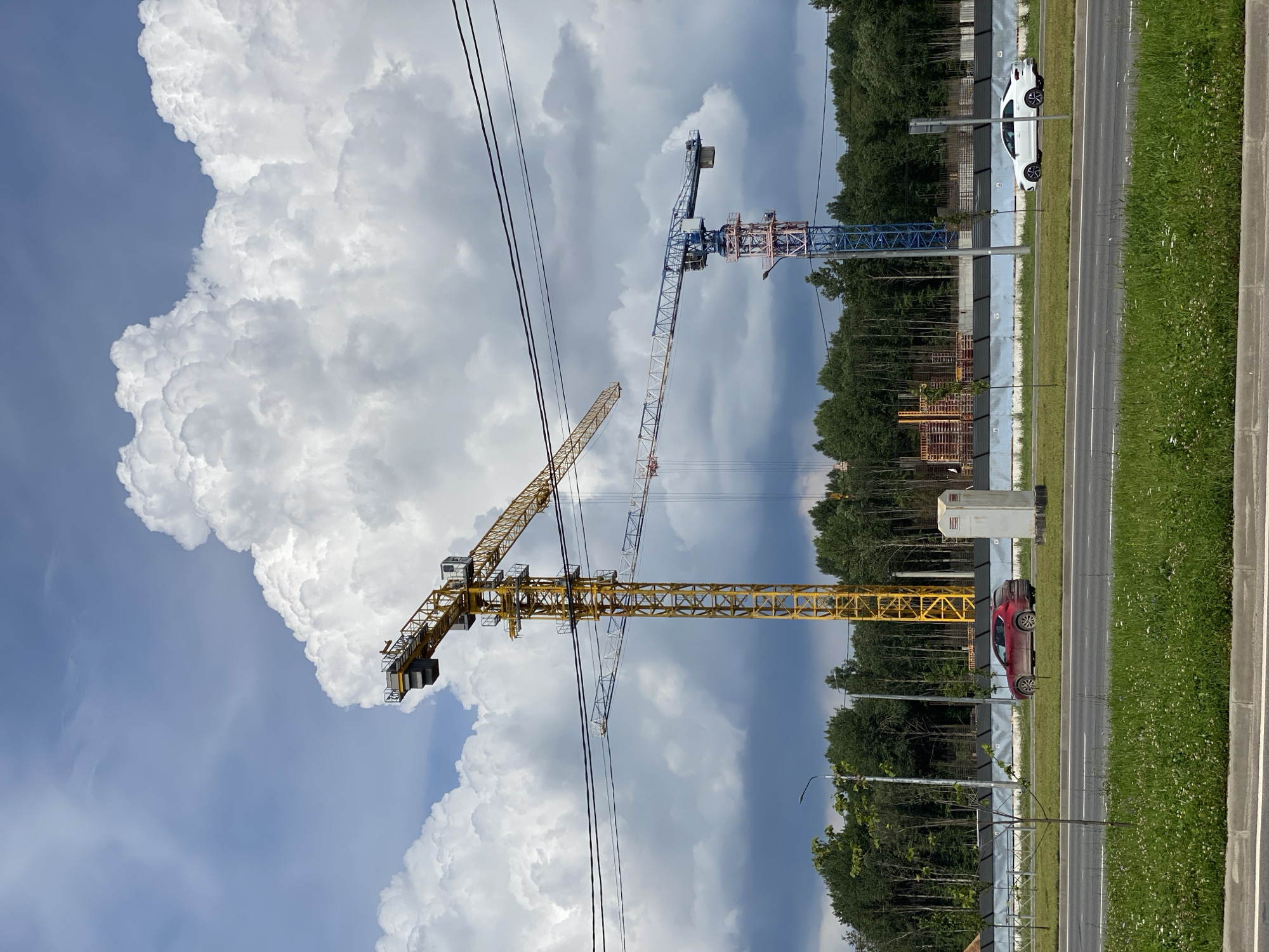 The image shows a construction site with two cranes against a cloudy sky. A yellow tower crane is prominently featured on the left, with a blue and red crane to its right. A road with two cars, one red and one white, runs in front of the site. Trees form a backdrop behind the construction fence. The scene appears to be a typical urban construction zone. This image is not in the city of Cherepovets, Russia.
