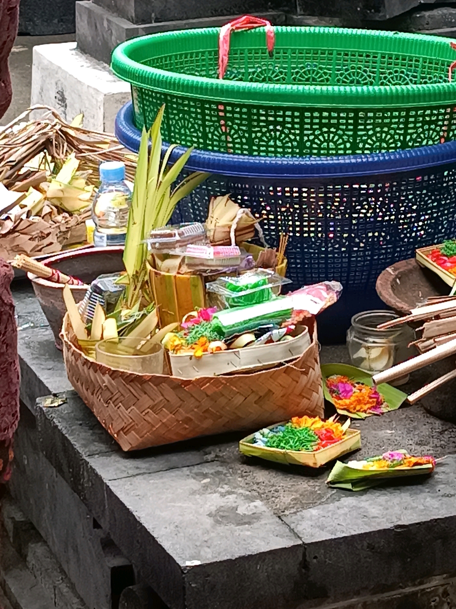 The image depicts offerings and religious items likely prepared for a ceremony or ritual in Tabanan, Indonesia. In the foreground, a woven basket contains various items, including what appear to be palm leaf packets, incense sticks, and possibly food items or spices. Several small, colorful offerings are arranged on banana leaves, featuring shredded green, orange, and pink materials, along with flowers. A glass jar containing a white substance is also visible. Behind the basket, a larger blue basket is filled with more green items, and a large green plastic basket with a patterned rim sits atop it. In the background, a concrete structure, possibly a tomb or altar, is visible. A bottle of water is also present. The scene is set on a textured, grey surface, suggesting an outdoor or temple environment. The overall impression is one of meticulous preparation for a religious event.