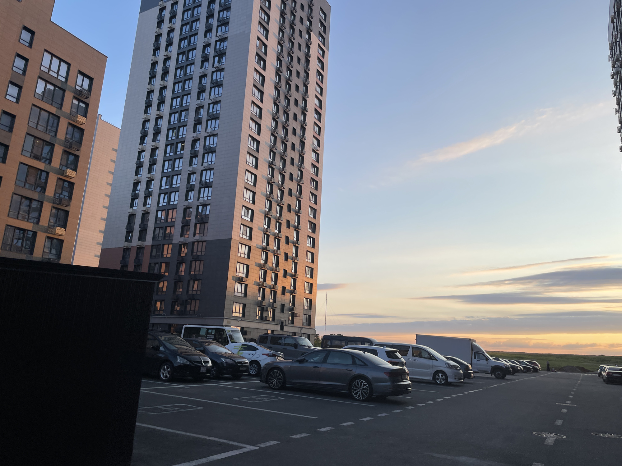 The image shows a view of a residential complex at sunset, likely in Stavropol, Russia. In the foreground, a parking lot is visible with several cars parked. The cars include a black sedan, a gray Audi sedan, a white van, and other vehicles. Behind the cars, a tall, modern apartment building with many windows is prominent. To the left of this building, another, shorter building with a reddish-brown facade is partially visible. The sky above is a soft blue with scattered, wispy clouds tinged with orange and yellow from the setting sun. The overall scene depicts an urban environment during the evening hours.