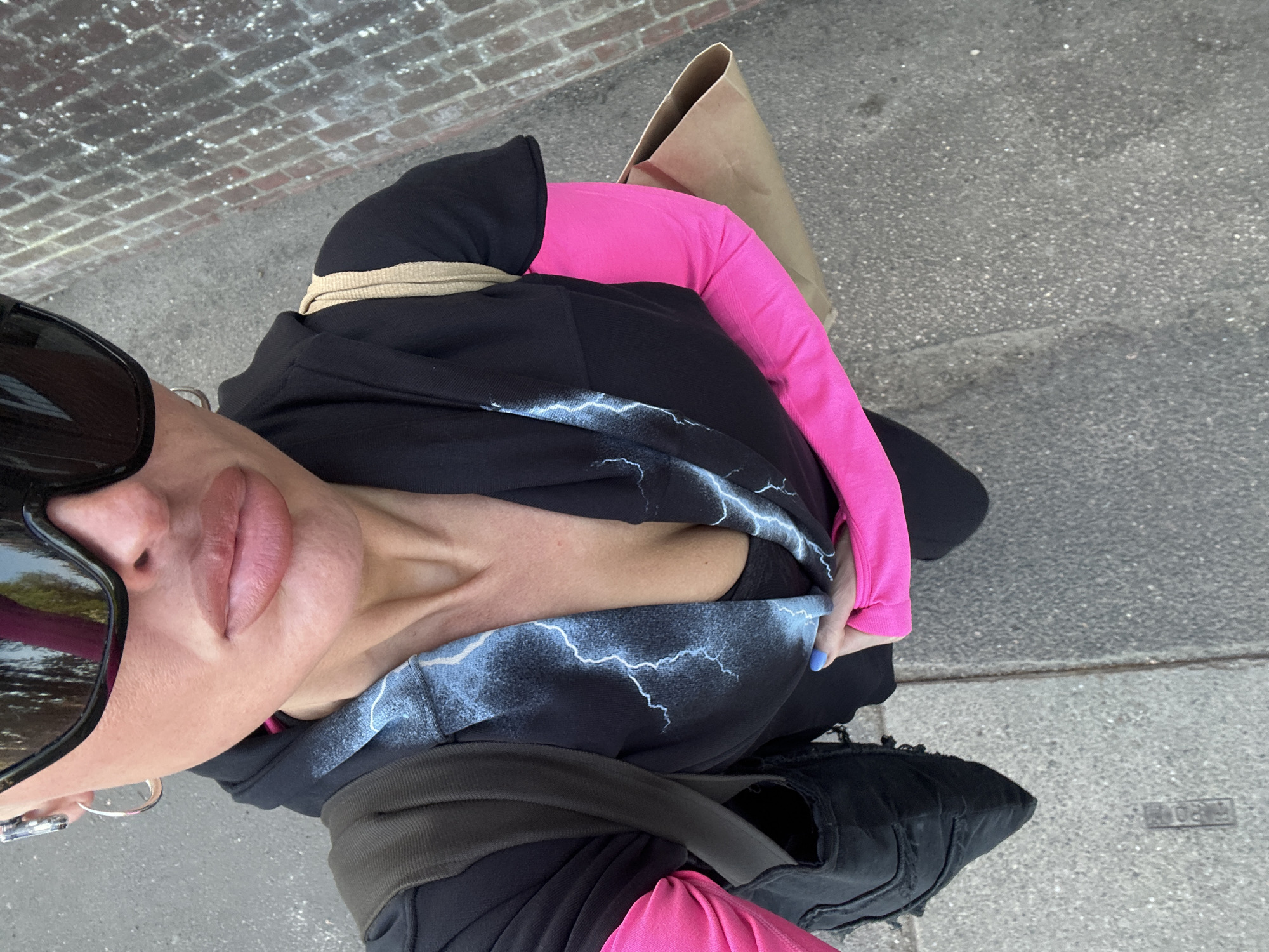 The image presents an angled, close-up outdoor shot of a person, likely a woman, looking towards the camera from an elevated perspective. The subject wears large, dark sunglasses that reflect the surroundings, and their lips appear full. They are dressed in a black hoodie adorned with a striking white and blue lightning bolt design across the chest. A bright, neon pink long-sleeved top is visible underneath the hoodie, particularly on the right arm. A light beige or khaki colored band is wrapped around the neck or collar area. The person's left hand, partially visible, sports blue nail polish and a silver ring or earring. Black trousers or leggings and black, textured shoes are also visible. A brown paper bag rests behind the person's right shoulder.
The setting is an outdoor urban or suburban environment, likely during daytime given the bright lighting and the use of sunglasses. The ground is grey asphalt or concrete, and a red brick wall is visible in the upper left background. A small, weathered metal plate or marking on the ground to the right of the person's feet contains indistinct raised text, possibly "P0R0" or a fragment of a word like "BORO". The overall impression is a casual, fashion-forward outfit worn in a street setting in Tunbridge Wells, United Kingdom.