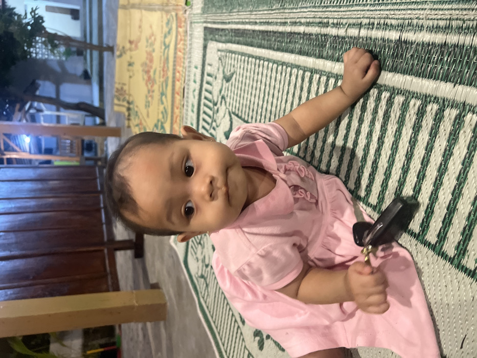 An infant, appearing to be of Asian ethnicity, lies prone on a green and white woven mat. The individual is dressed in a short-sleeved pink collared dress and holds a dark-colored object, possibly a key fob with a metallic ring, in their right hand. The infant's head is turned to the left, and lips are slightly puckered. In the background, a light brown wooden pillar is present on the left, with a dark wooden screen behind it. To the right, a yellow-patterned mat, a tree, and a light-colored building with visible artificial lighting are discernible. The ground beneath the mats appears to be a paved surface. The scene takes place in Mojorejo, Indonesia, under artificial illumination, indicating evening or night conditions.