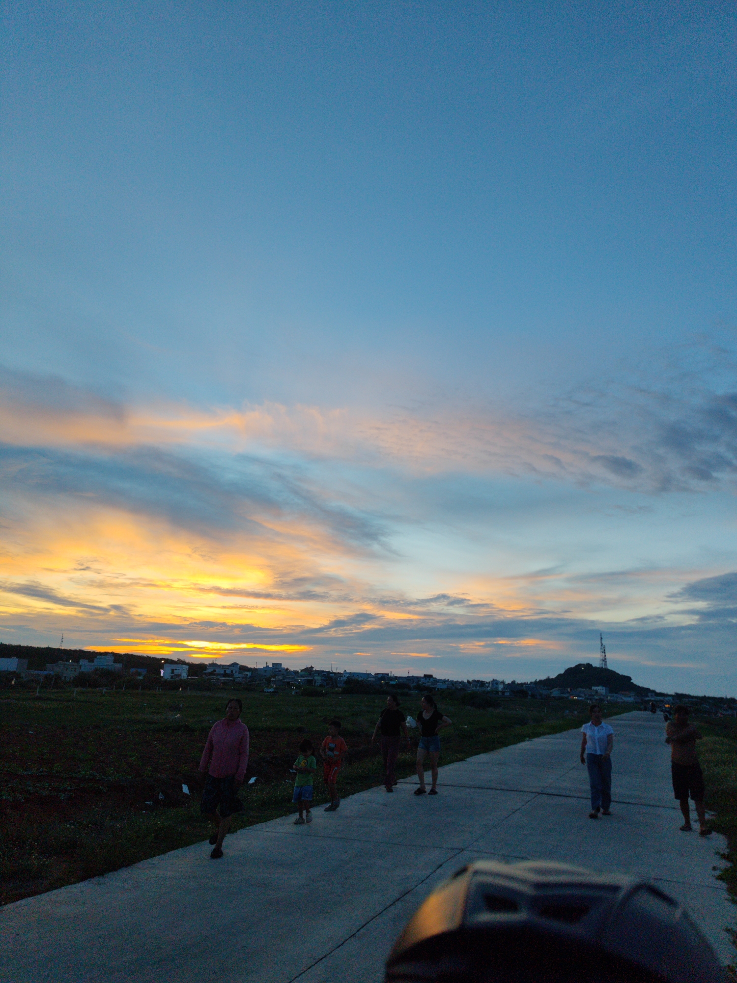 The image depicts a group of people walking along a concrete path in Phú Quý Special Administrative Region, Vietnam, at sunset. The sky is a mix of orange, yellow, blue, and gray, with the sun setting behind a line of buildings and foliage on the horizon. A hill with a tall transmission tower is visible in the distance on the right. A woman and two children walk on the left side of the path, while another group of three people walks further ahead, and a shirtless man stands on the right. The bottom of the image shows the top of a dark object, possibly a vehicle's dashboard.