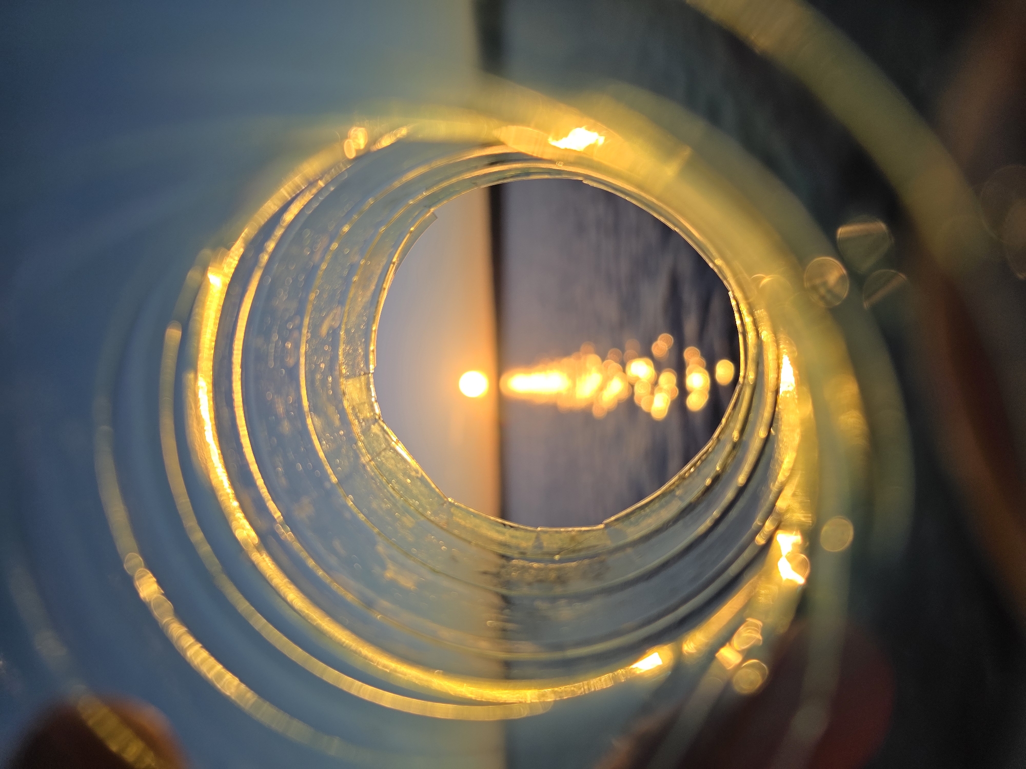 The image shows a sunset over water as seen through the inside of a translucent tube. The tube is mostly out of focus, creating blurry rings. The sunset is centered in the opening of the tube, with a bright sun reflecting off the water's surface. The image appears to have been taken near Chaikovsky, Russia, based on the available data. No people are directly visible. The interaction is between the sunlight, the water, and the light's passage through the tube. The environment is a body of water with a setting sun.