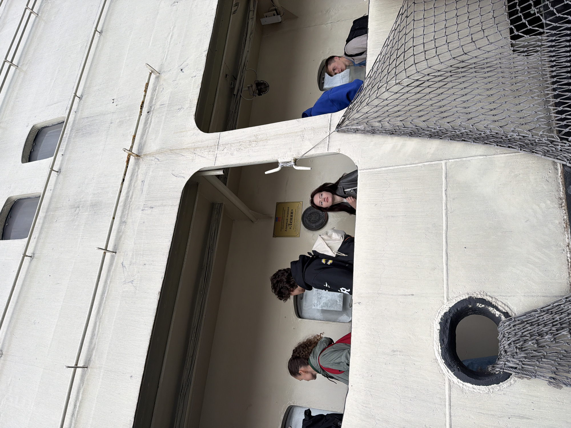 Four individuals stand on an elevated walkway of a large, white, industrial-looking structure, possibly a ship or a building with similar architecture. The structure features windows and railings. A netted barrier is prominent in the foreground on the right side of the image. The individuals appear to be looking in different directions. The person on the far left is a woman with curly hair wearing a green jacket and a red backpack. Next to her is a young man with dark curly hair wearing a black hoodie and a light-colored backpack. To the right of him, a young woman with long dark hair wears a grey jacket. On the far right, a young man with short blonde hair wears a beige backpack and a grey top. A sign with Cyrillic text, including "АТОМНЫЙ ЛЕДОКОЛ «ЛЕНИН»" (Atomic Icebreaker "Lenin"), is visible on the wall behind the people. This indicates the location is likely the Atomnyy Ledokol "Lenin" museum ship in Murmansk, Russia.