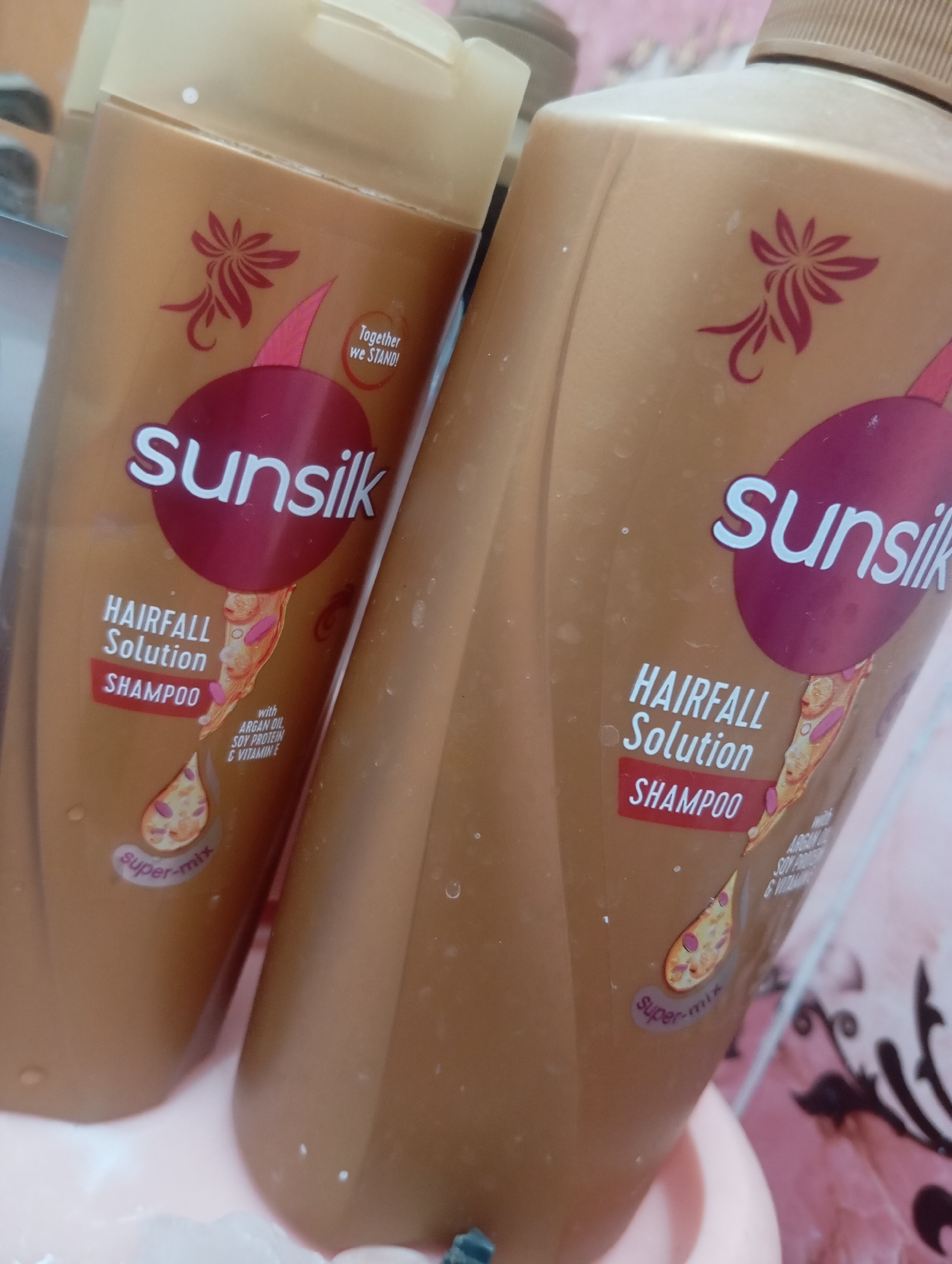 Two bottles of Sunsilk Hairfall Solution Shampoo are displayed in a close-up shot. The bottles are a golden-brown color with maroon labels. The left bottle is slightly out of focus. Both bottles feature the Sunsilk logo and text indicating "Hairfall Solution Shampoo". Below this, a graphic depicts a drop containing argan oil, soy protein, and vitamin E. A small circular label at the bottom reads "super-mix". The bottles appear to be placed on a pink surface, with a hint of a patterned background and what looks like a mirror reflecting the tops of the bottles. The scene is indoors, but the specific location within a city is unknown.