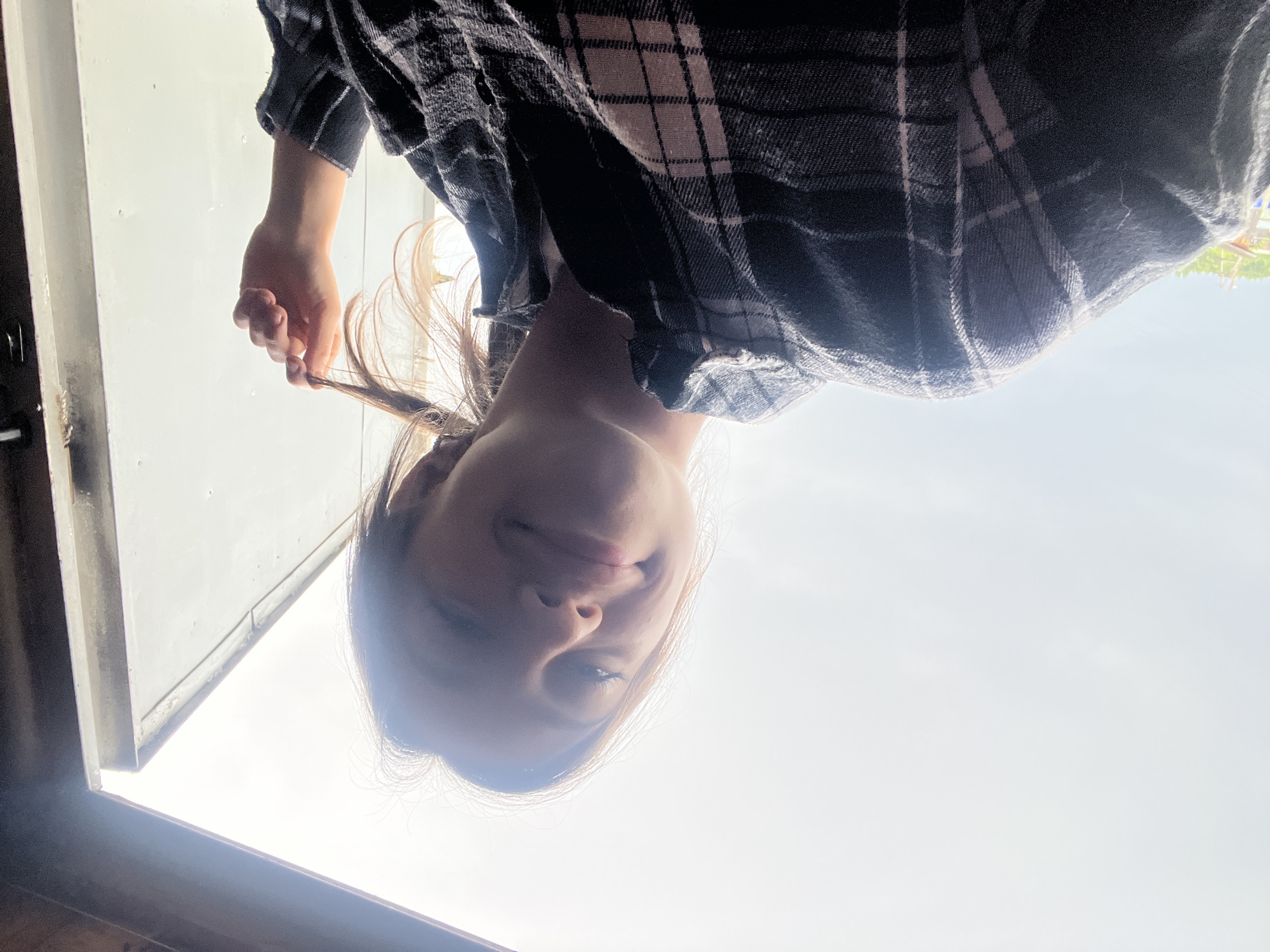 A young woman is captured in a low-angle selfie, outdoors in what appears to be a rural or semi-rural setting. She is wearing a dark plaid shirt. Her hair is light brown, and she is twirling a section of it with her right hand. The background is dominated by a bright, overcast sky and a white, slightly weathered structure, possibly a wall or a door, with a dark frame. The lighting suggests it is daytime. No specific landmarks or context are provided to pinpoint a location within the city of Kemul, Russia.
