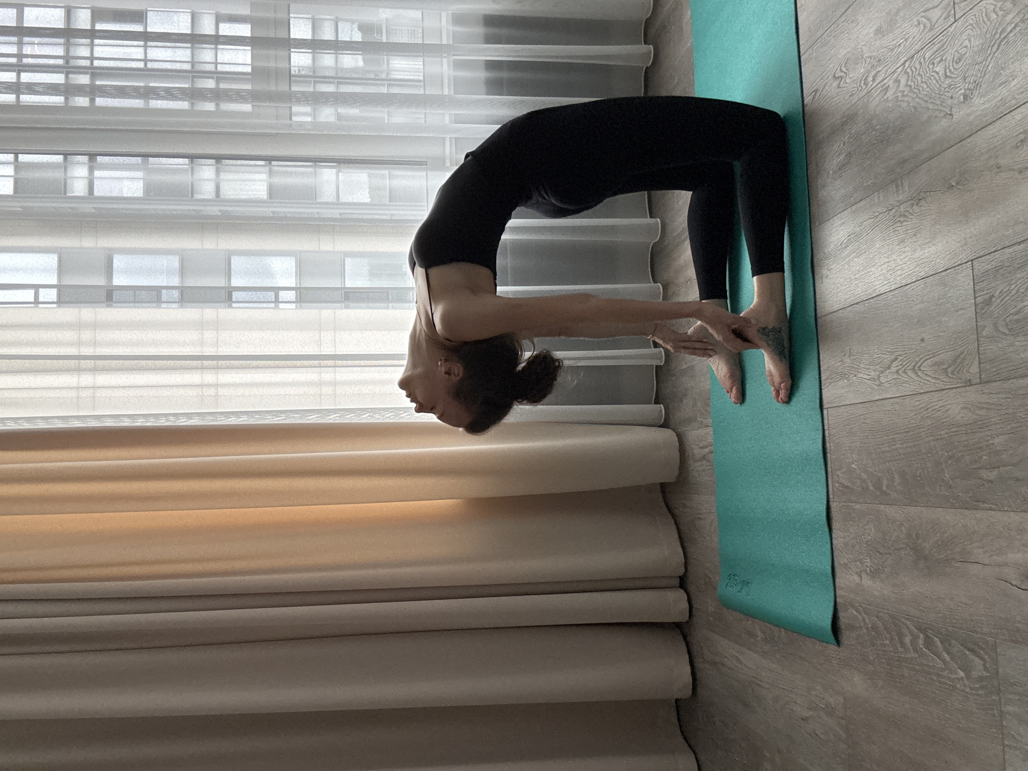 A woman is performing a yoga pose, specifically a backbend, on a teal yoga mat. She is wearing black leggings and a black tank top. Her body is arched backward, with her hands resting on the mat behind her feet. Her head is tilted back, and her hair is pulled up in a bun. The yoga mat is placed on a light-colored wooden floor. In the background, there are sheer white curtains covering windows, which show a blurry view of a building. To the left of the yoga mat are folded, light brown curtains. The lighting appears to be natural, suggesting daytime. The scene is indoors, likely in a home or studio. The overall mood is one of focused effort and physical exertion. No visible text is present in the image.