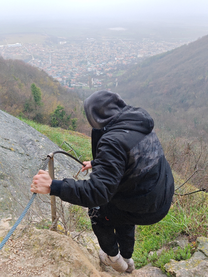 A person of indeterminate gender is positioned on a steep, rocky Via Ferrata route overlooking the city of Shumen, Bulgaria. The individual wears a dark hooded jacket, dark pants, and light-colored athletic shoes. Their left hand grips a thick steel safety cable, while their right hand holds a silver carabiner near the cable. The immediate foreground consists of rugged, light-colored rock and sparse green vegetation. The background features a panoramic view of Shumen city nestled in a valley, characterized by a dense arrangement of buildings with varied roof colors and discernible road networks. Surrounding hillsides are partially covered with bare deciduous trees and some evergreen patches, indicating a cool season. The sky is uniformly overcast, creating a hazy atmosphere that softens the distant urban landscape.