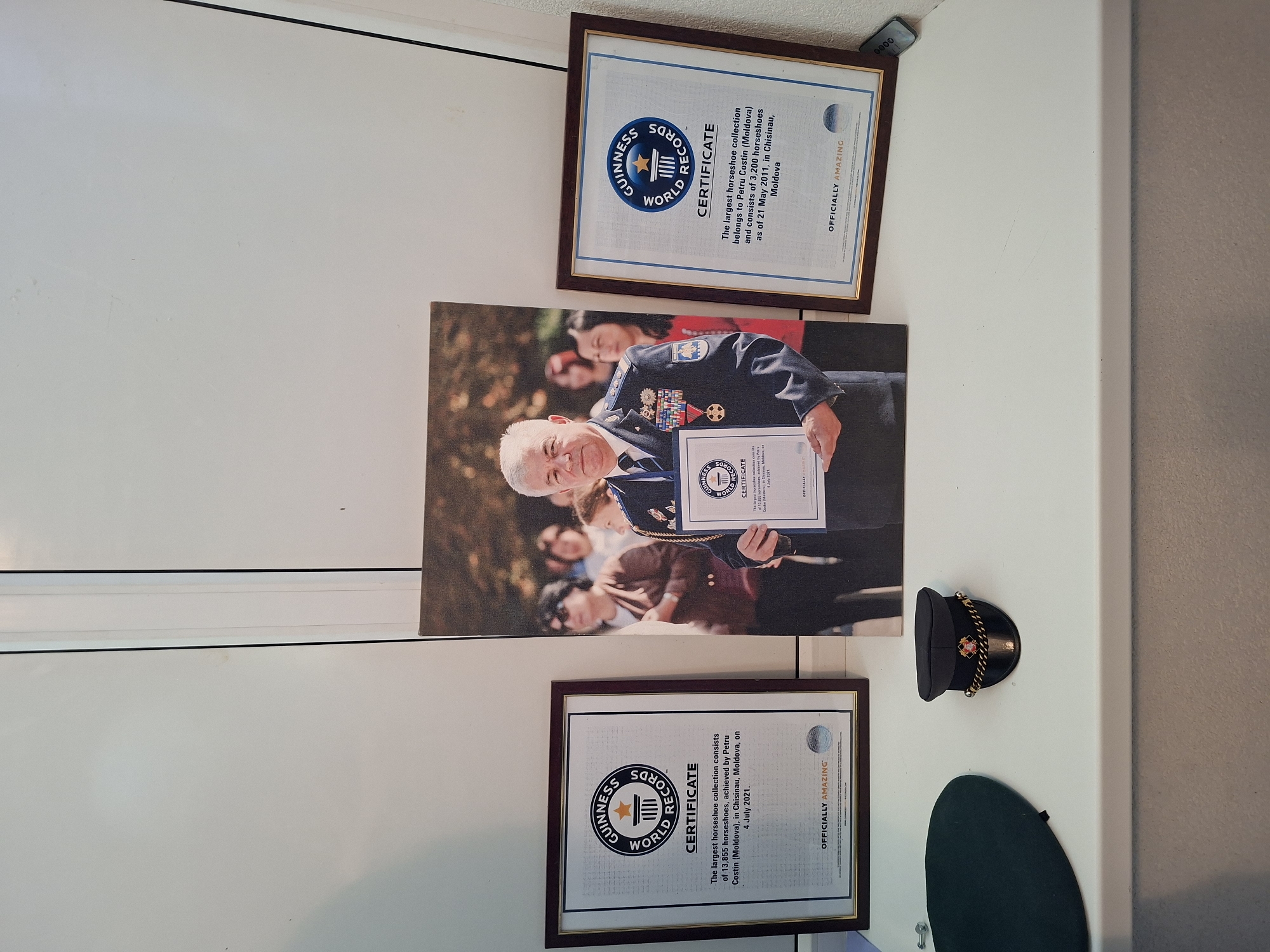 The image depicts a tabletop display featuring a framed Guinness World Records certificate on the left, a large framed photograph of a man in uniform holding a certificate in the center, and another framed Guinness World Records certificate on the right. A dark green beret is visible on the far left, and a small peaked cap with gold trim sits between the left certificate and the photograph. The certificates are official documents from Guinness World Records, one dated 4 July 2021, and the other 21 May 2011, both related to horseshoe collections achieved by Petru Costin from Chisinau, Moldova. The photograph shows a man with medals and insignia on his uniform, holding a certificate. The background appears to be a white wall with a vertical seam. The scene is set on a white surface, possibly a shelf or windowsill, in what is likely a domestic or personal display area in Ialoveni, Moldova.